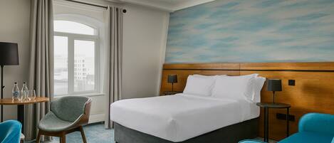 A hotel room with a bed, a chair, a small table with a lamp, and a view of the city through a window.