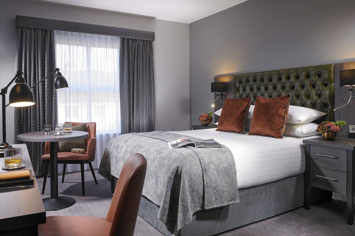 A modern hotel room with a large bed, a desk with a chair, a round table, and a window with curtains.