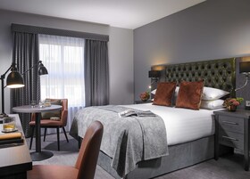 A modern hotel room with a large bed, a desk with a chair, a round table, and a window with curtains.