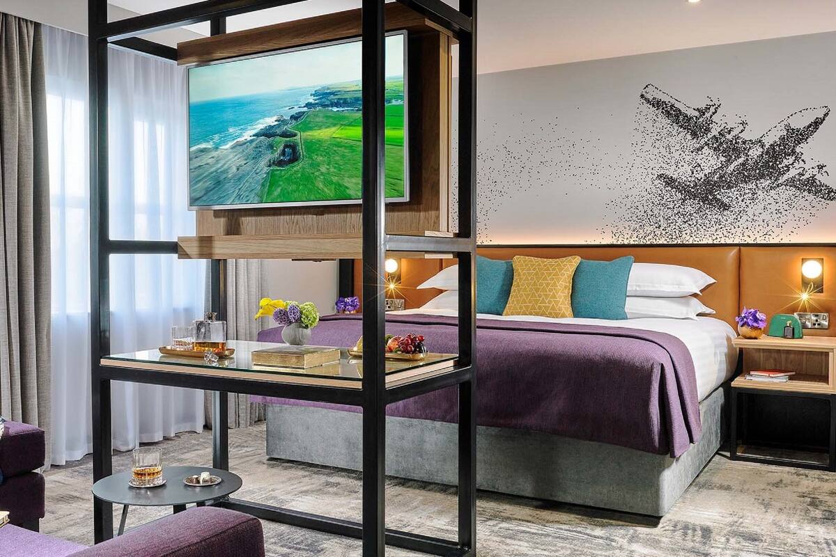 A hotel room with a large TV, a bed with a purple and white bedspread, a nightstand with a lamp, and a view of the ocean.