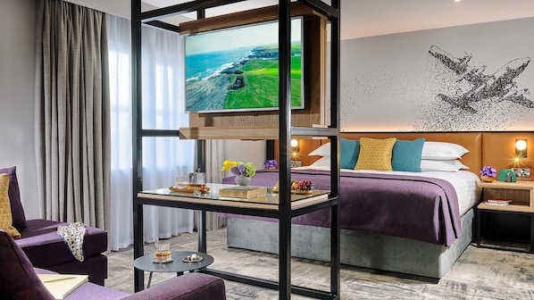 A hotel room with a large TV, a bed with a purple and white bedspread, a nightstand with a lamp, and a view of the ocean. - Shannon Springs Hotel (Shannon)