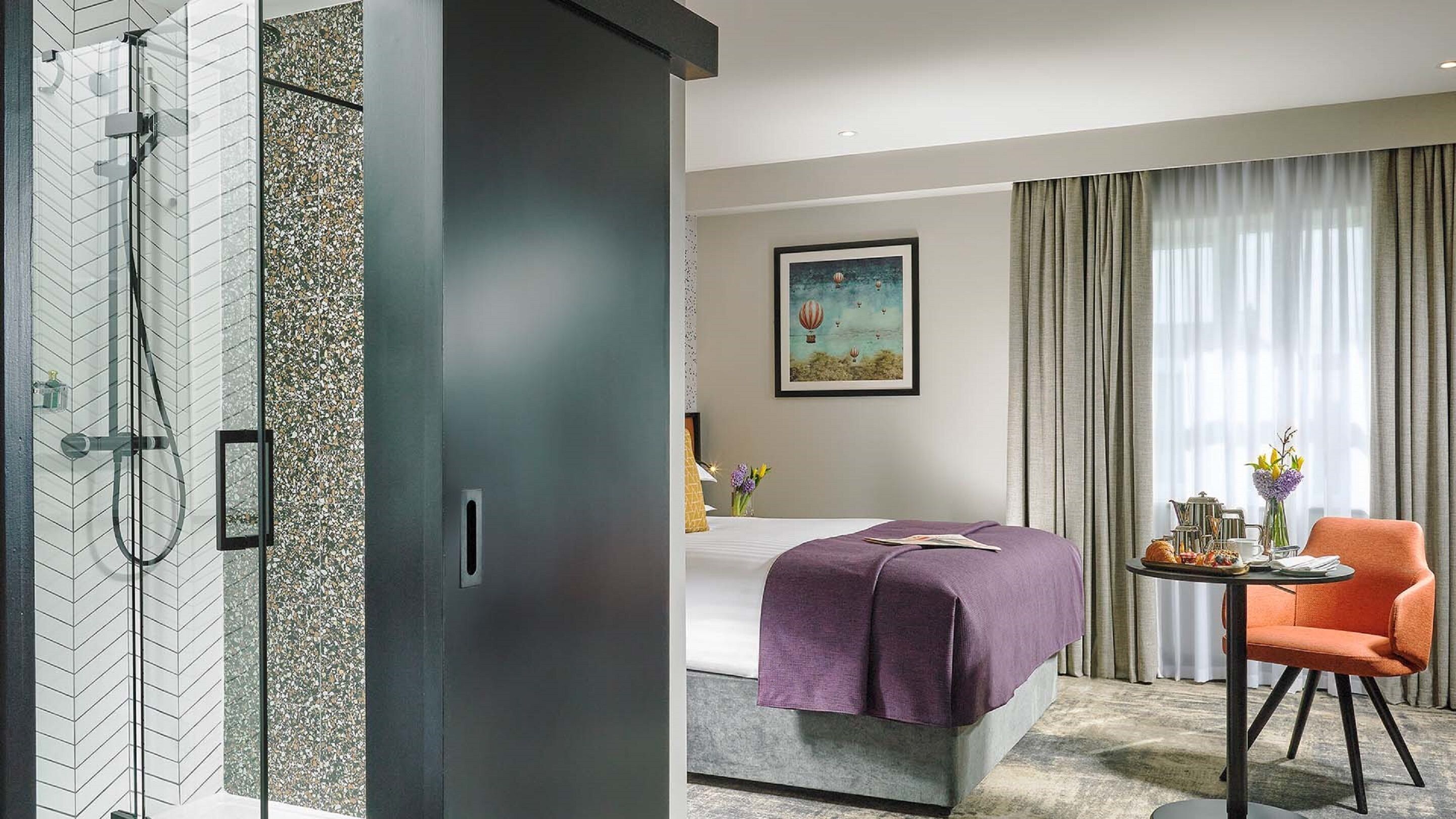 Una habitación de hotel moderna con ducha, una cama con una manta morada, una mesa de comedor con productos para el desayuno y una silla.