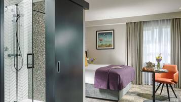 Una habitación de hotel moderna con ducha, una cama con una manta morada, una mesa de comedor con productos para el desayuno y una silla.