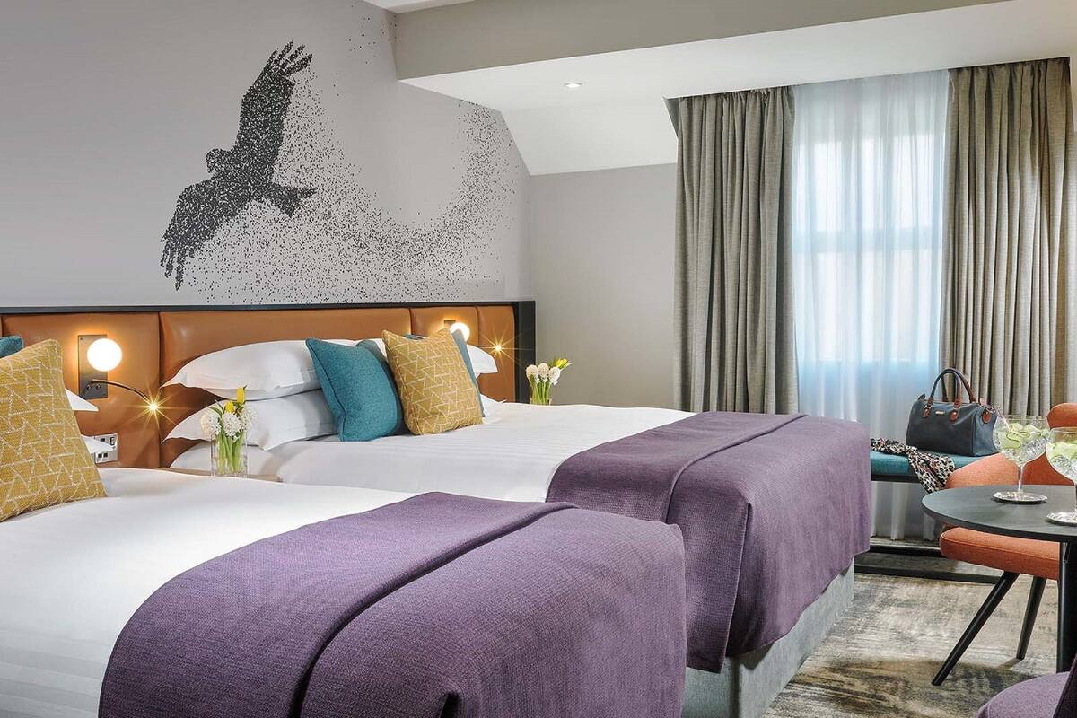 A hotel room with two beds, a large wall mural, a desk with a chair, and a view of the outdoors.