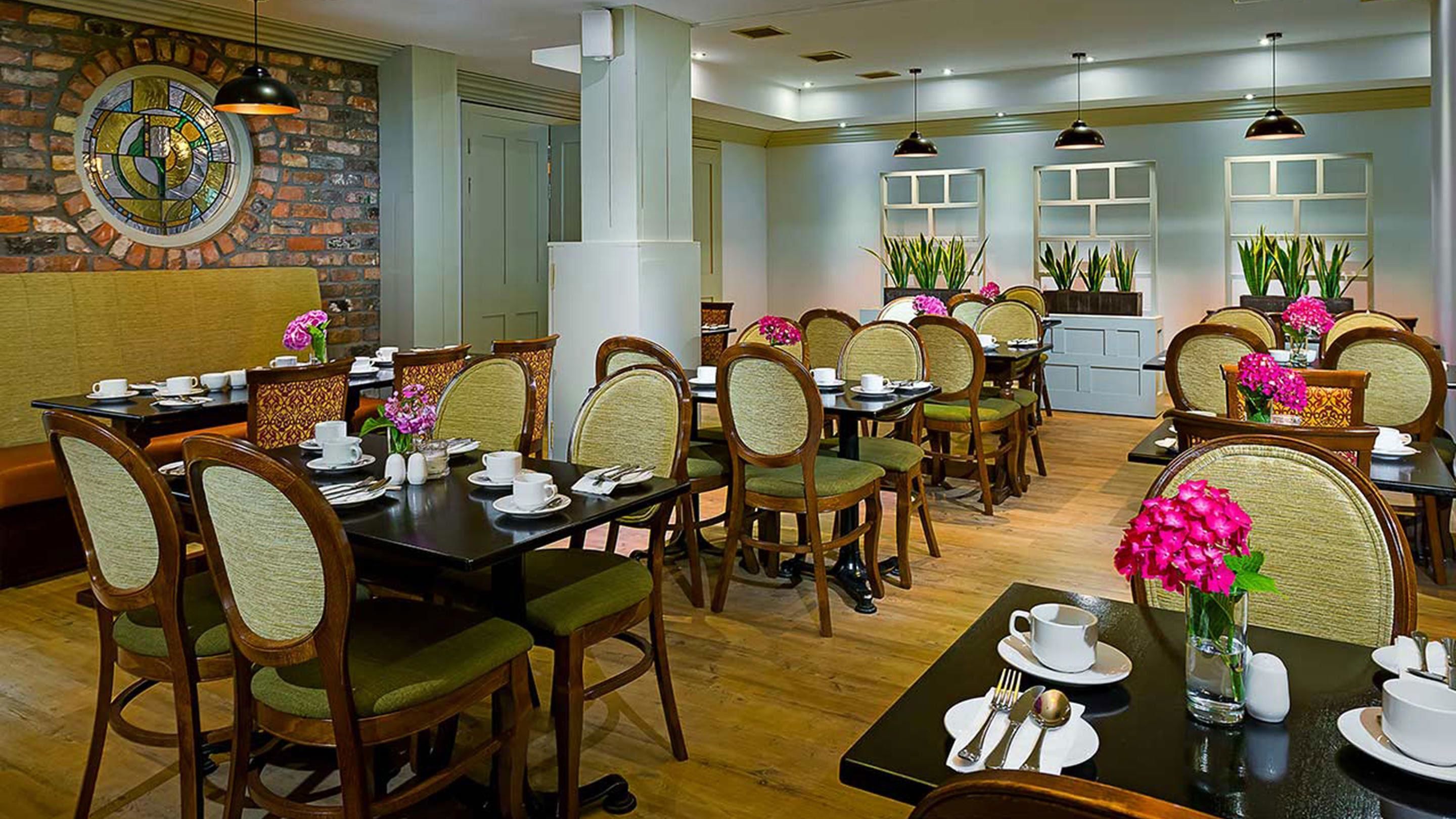 A spacious dining area with multiple tables and chairs, a brick wall with a stained glass window, and potted plants.