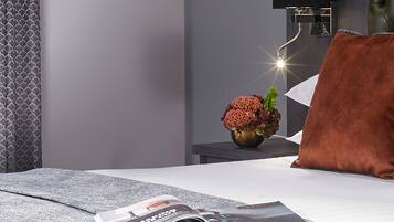 Un dormitorio moderno con una cama, una revista, un florero con flores y una mesita de noche con una lámpara.