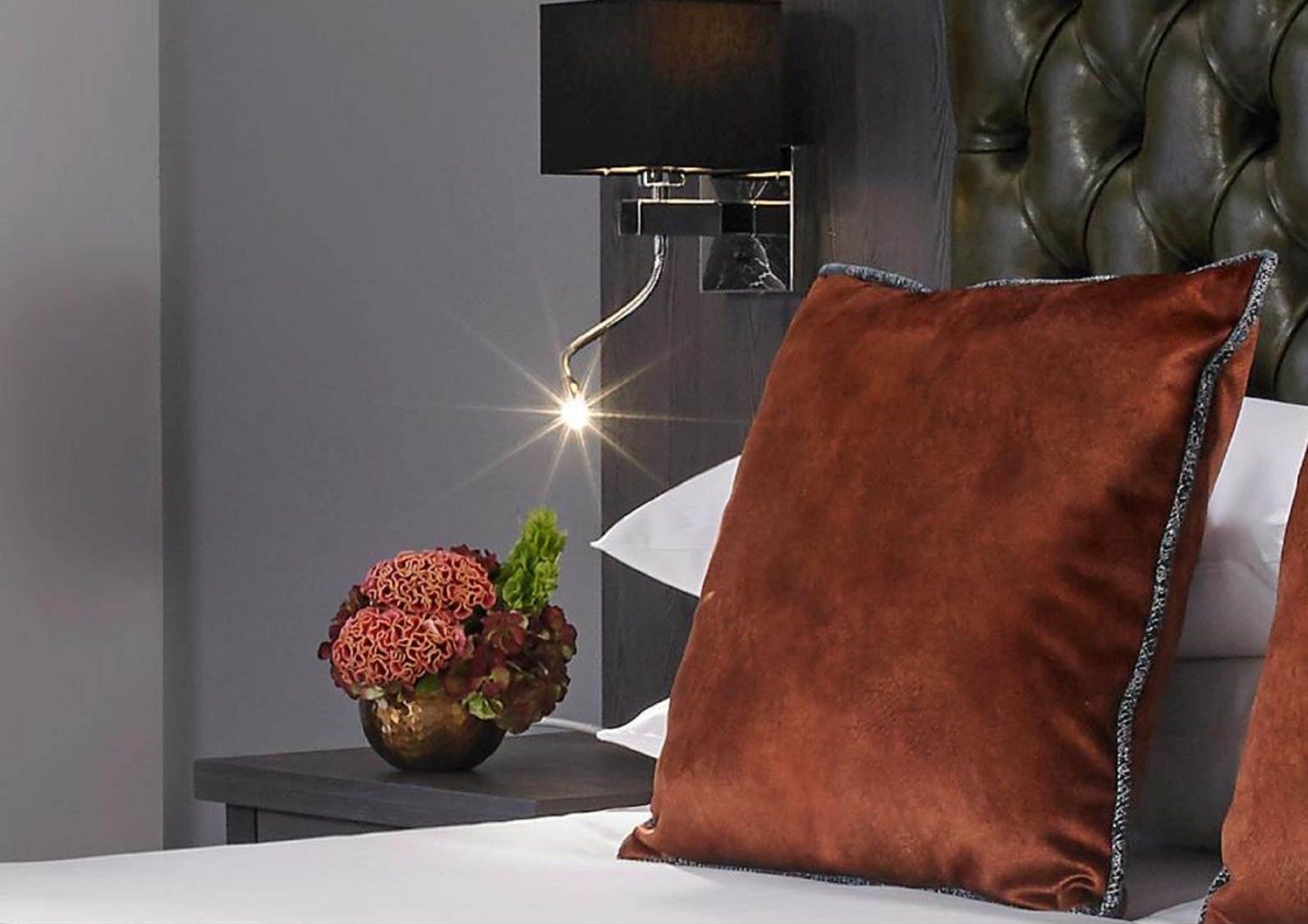 Un dormitorio con una cama, una mesita de noche, una lámpara y un florero decorativo con flores.