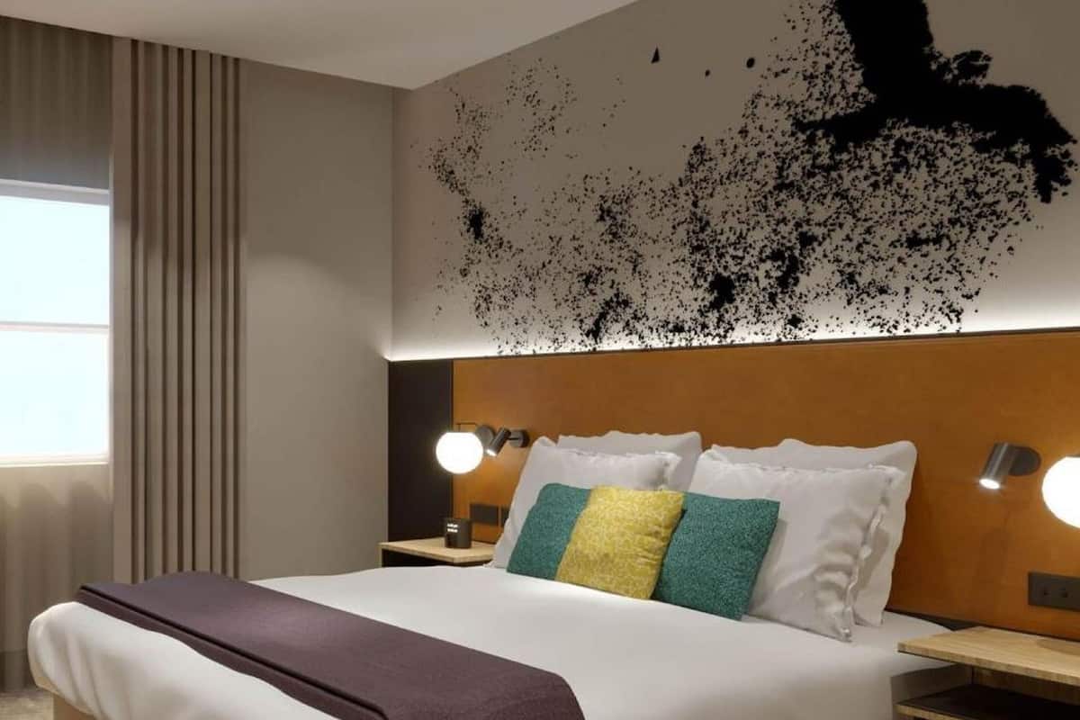 A hotel room with a large bed, two bedside tables, a wall-mounted light fixture, and a window with curtains.