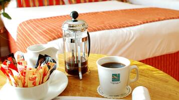 Una habitación de hotel con una cama, una mesita de centro con una cafetera, una taza de café, un bol de galletas y una revista.