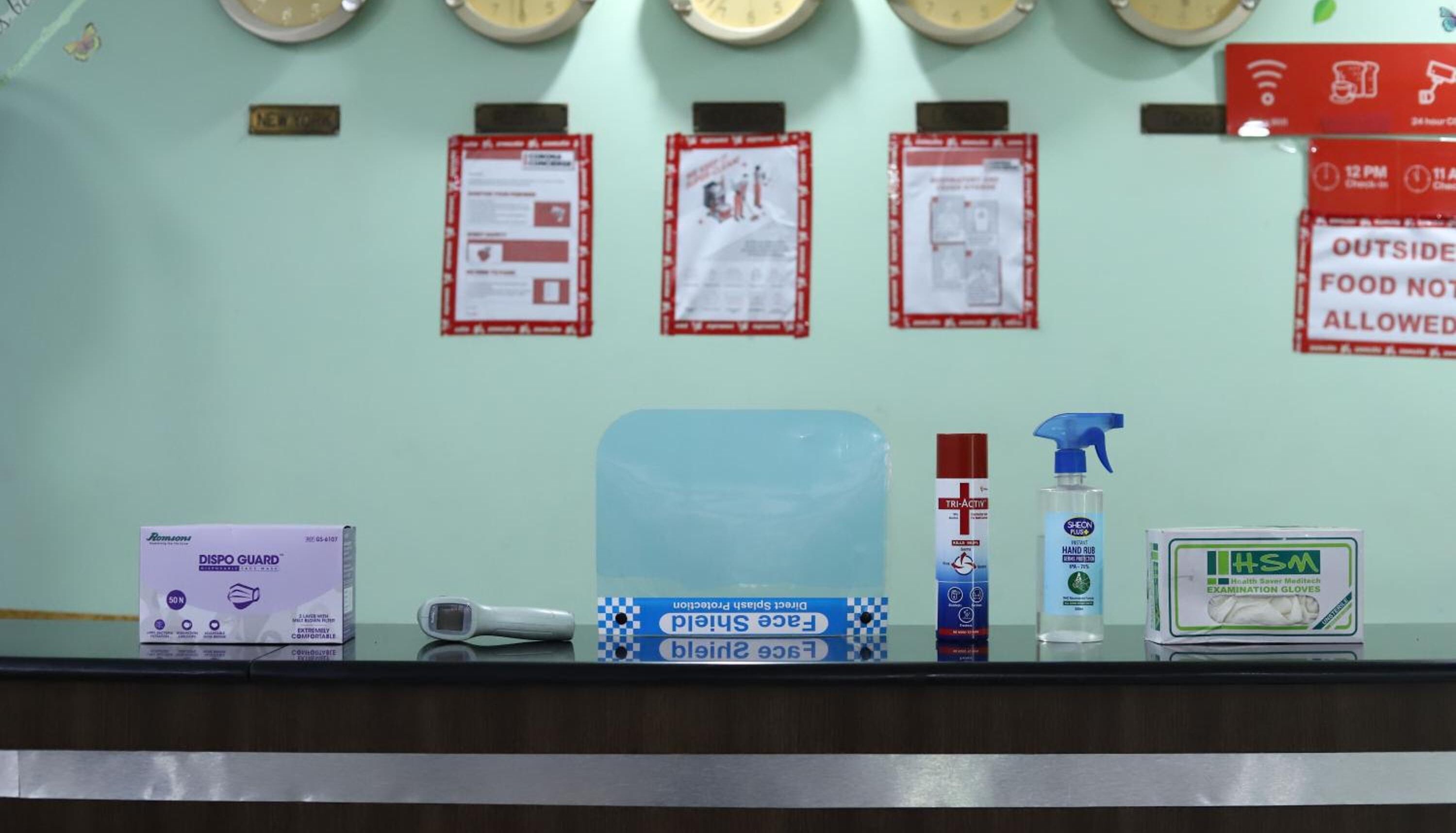 Mostrador de recepción con un reloj digital, dispensador de gel hidroalcohólico, caja con protectores faciales y dispensador de gel antibacterial de triple acción.