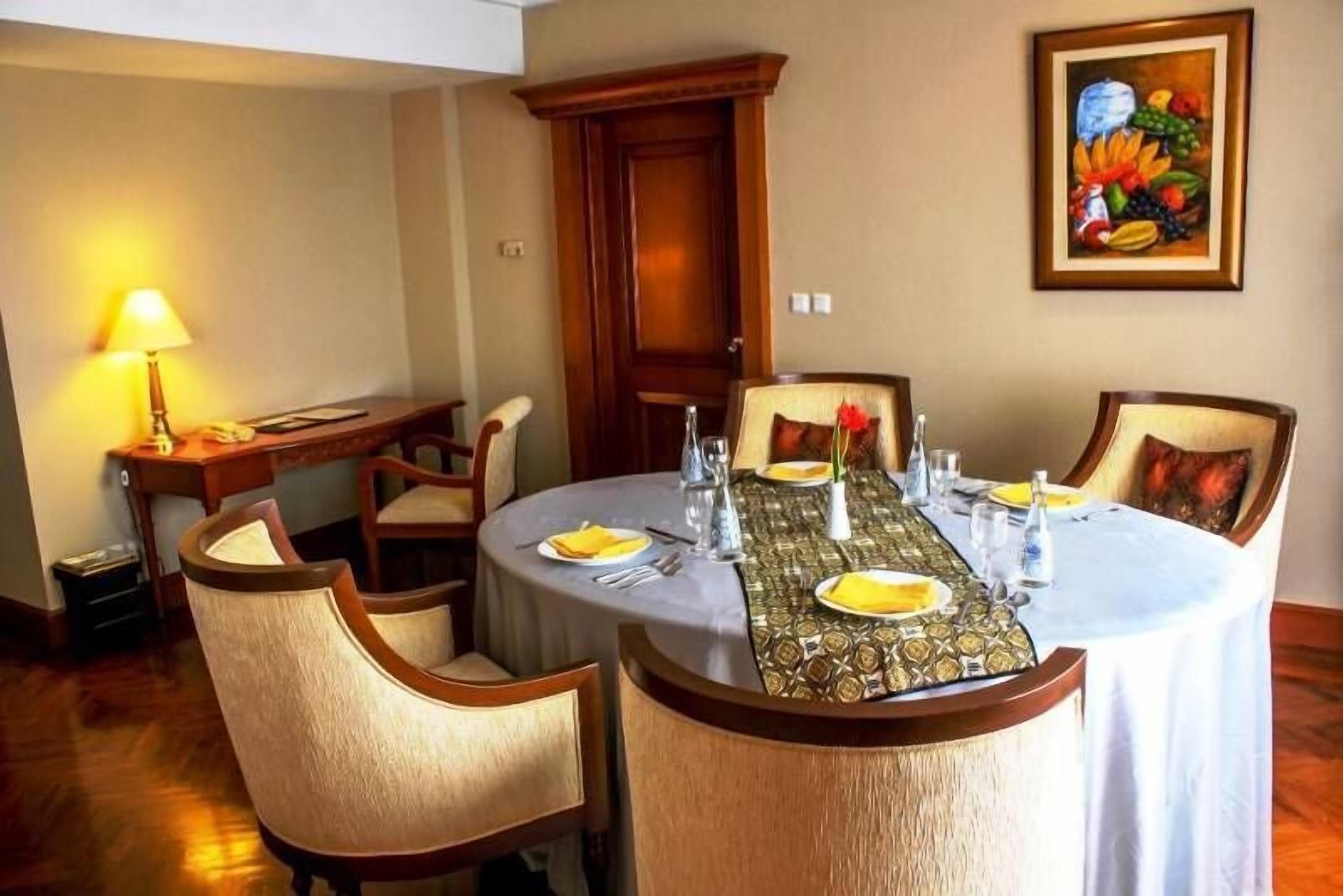 Un comedor con una mesa redonda dispuesta para cuatro personas, rodeada de sillas tapizadas. Un bodegón con frutas cuelga en la pared, y una lámpara está sobre una mesita auxiliar.