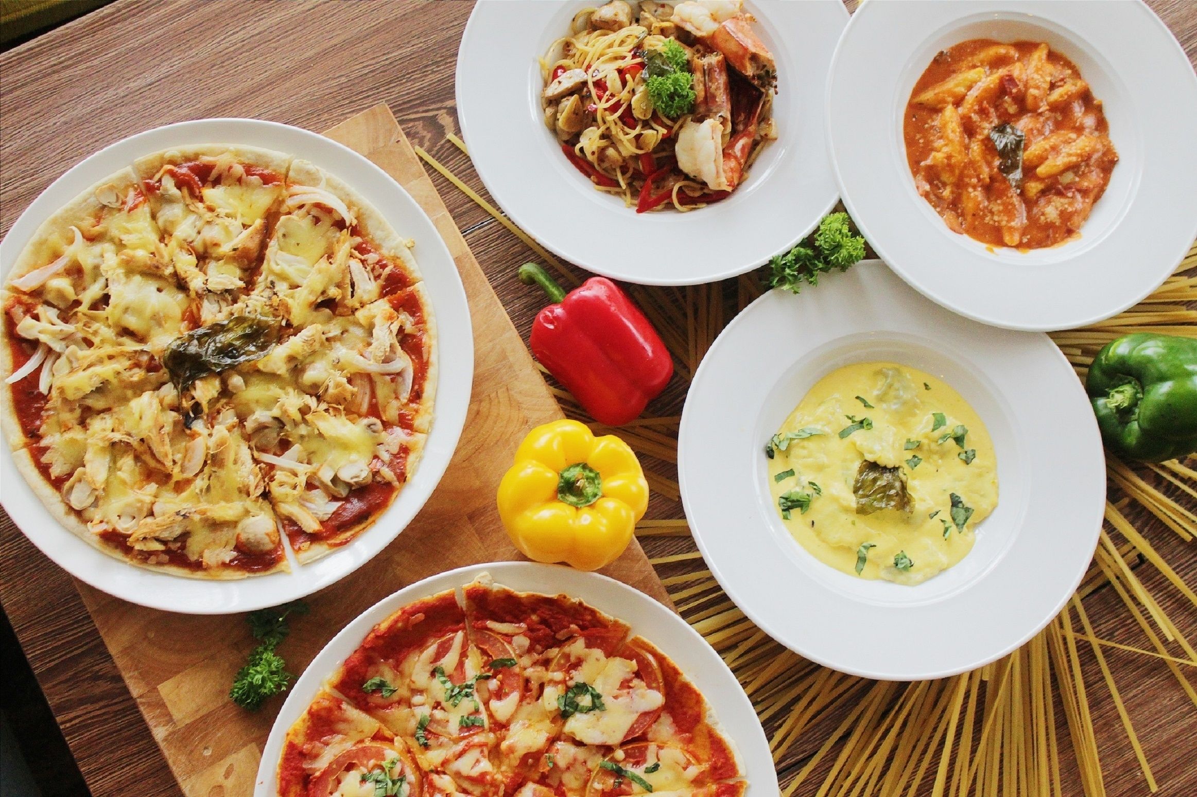 Una mesa con cuatro platos de comida, incluyendo pizza y pastas, rodeada de verduras y espagueti.