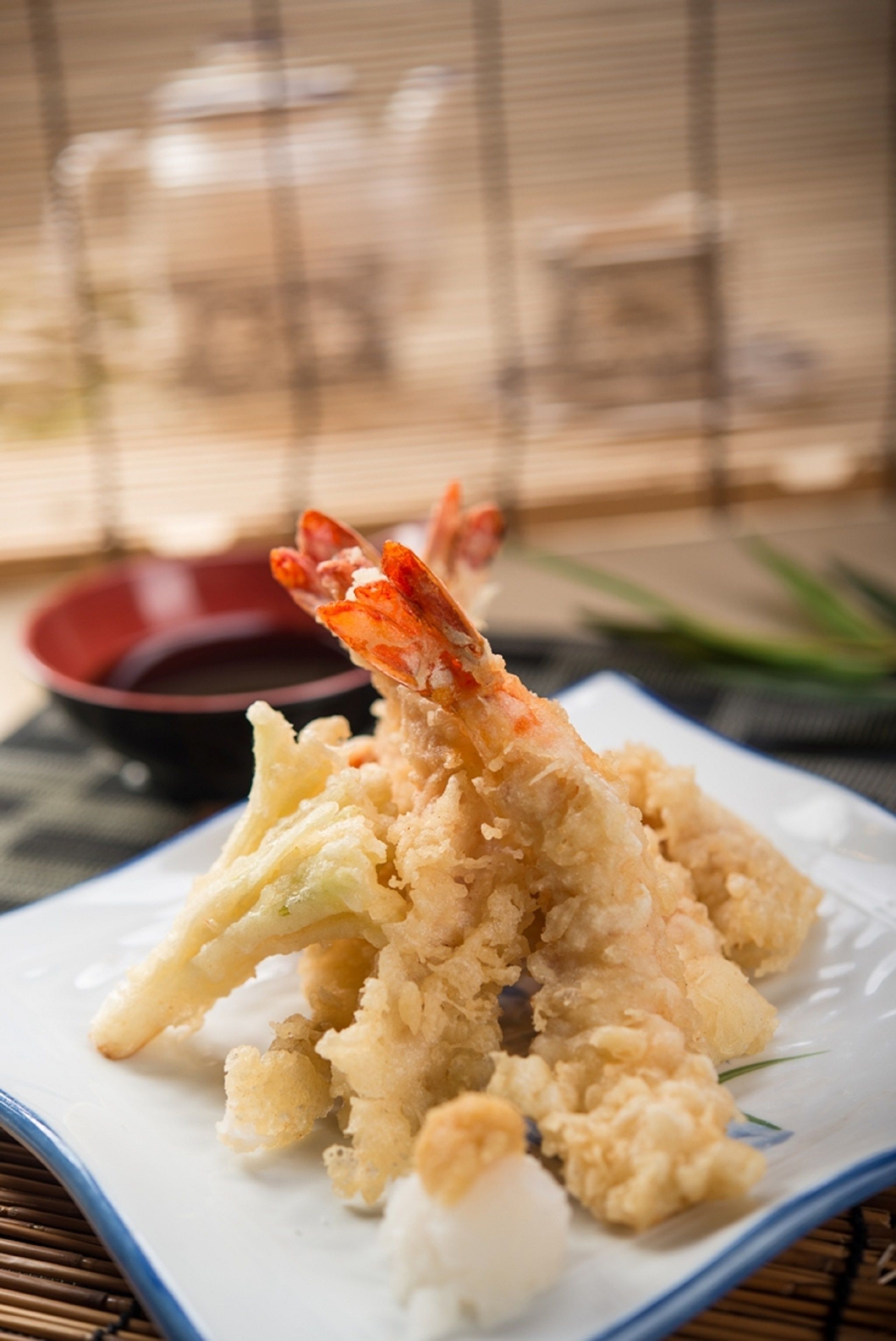 A plate of tempura with a shrimp on a white plate with a blue rim, placed on a table with a bamboo mat underneath.