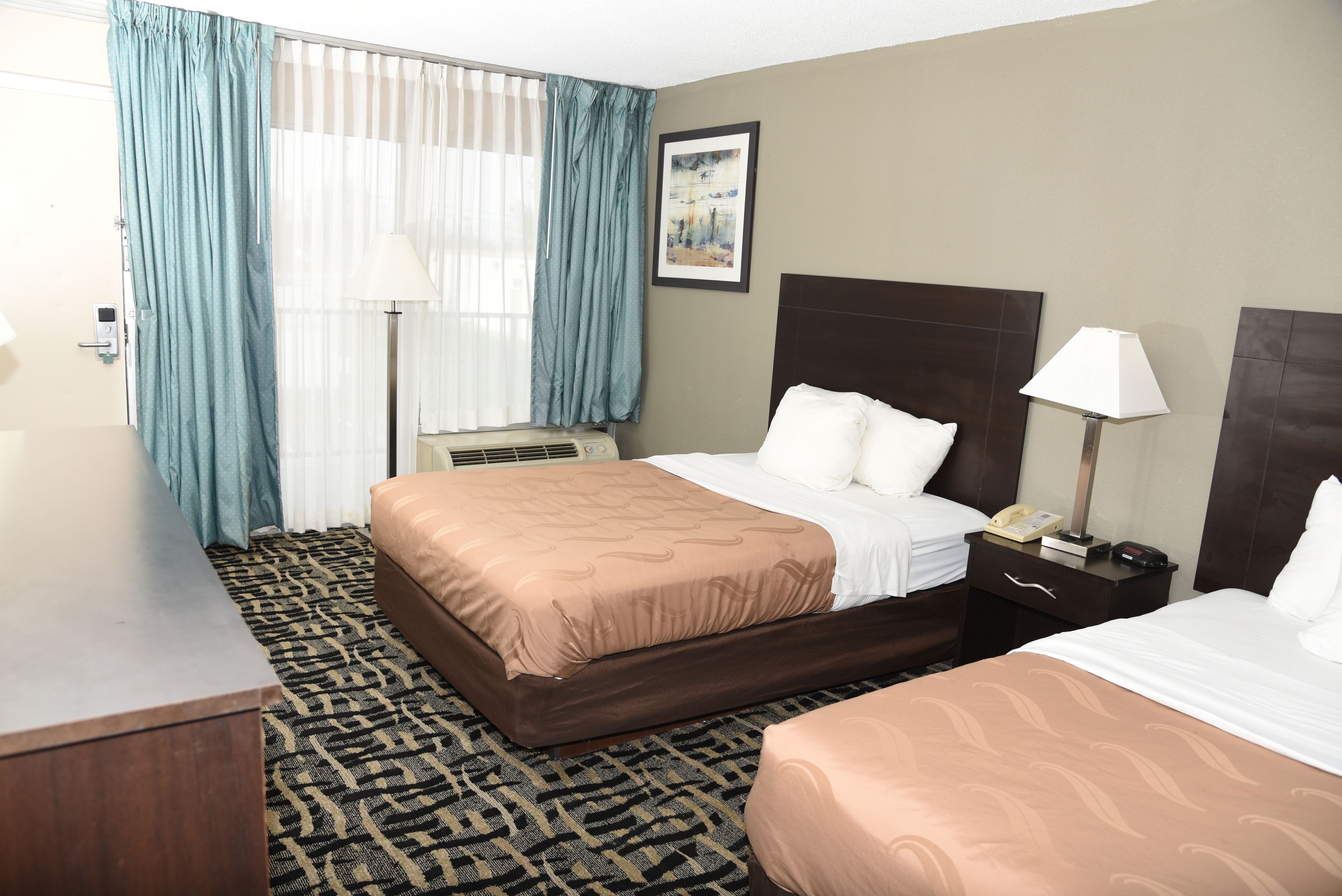 A hotel room with a large bed, a smaller bed, a desk, a chair, a lamp, and a painting on the wall.