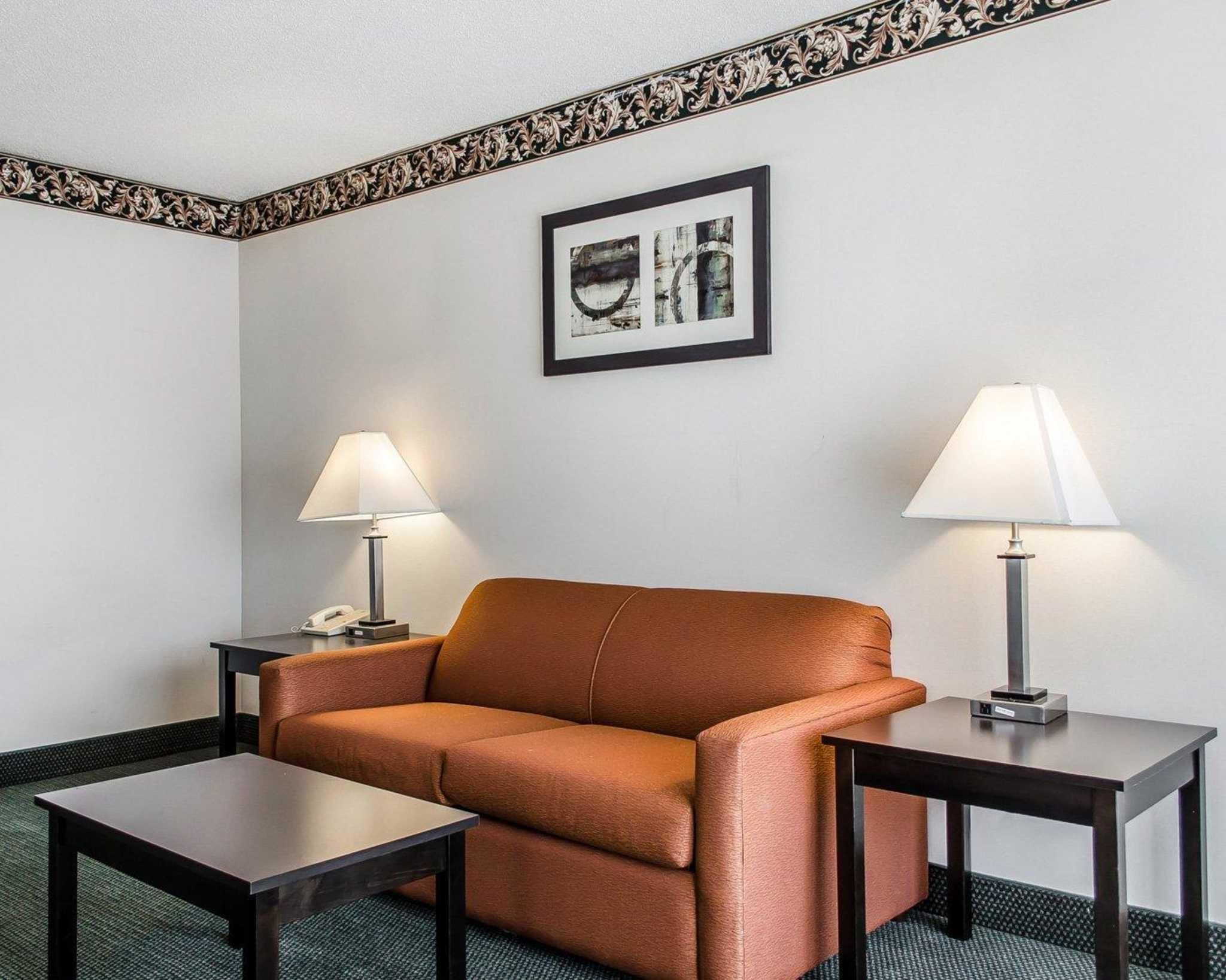 A hotel room with a brown leather sofa, two bedside tables with lamps, a framed picture on the wall, and a carpeted floor.