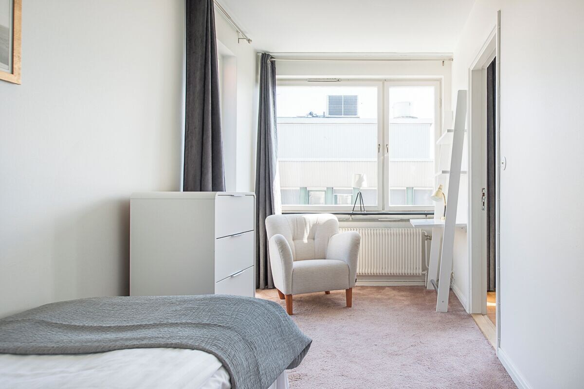 A bedroom with a bed, a chair, a desk, and a window with a view of buildings.