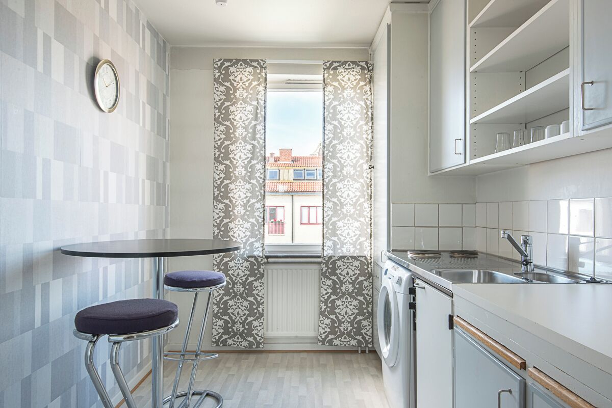 A compact kitchen with a washing machine, a sink, and a window with patterned curtains.
