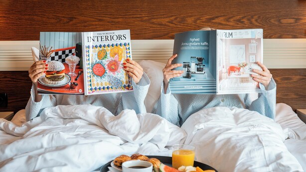 Two people reading books on a bed with breakfast. - ProfilHotels Grand Kristianstad (Kristianstad)