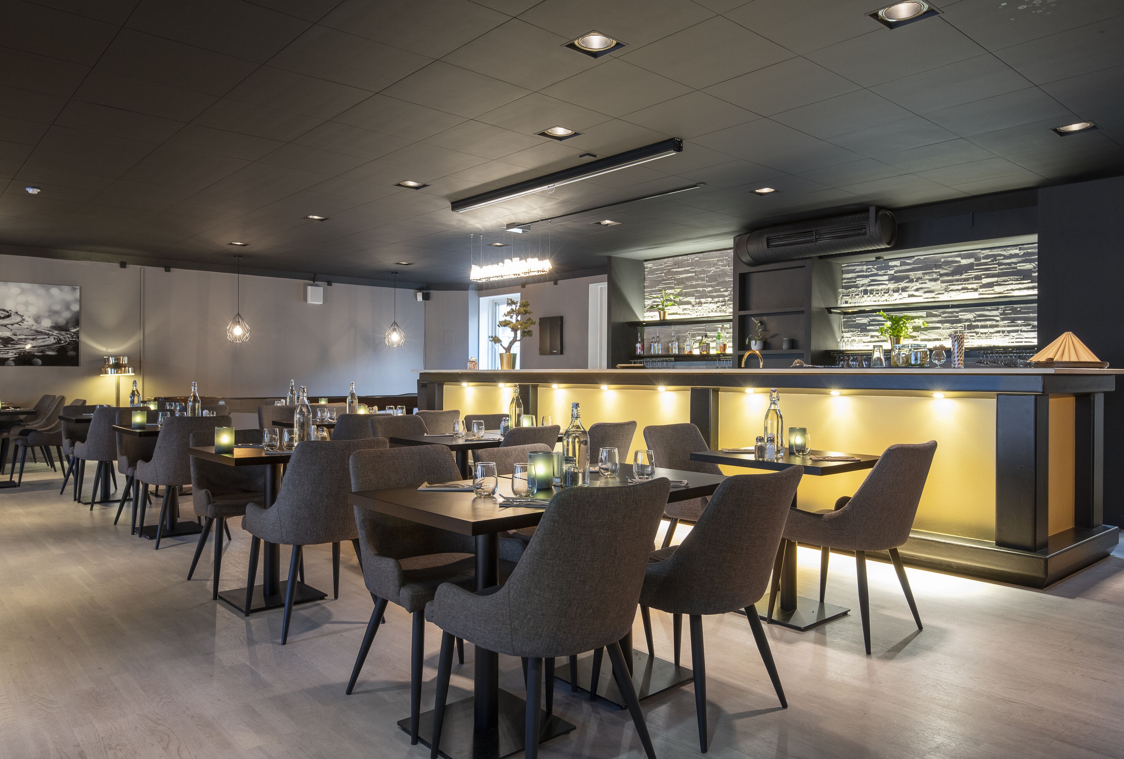 A modern restaurant with multiple dining tables and chairs, a bar area, and a kitchen visible through a large window.