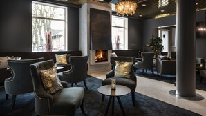 A modern lounge area with dark-toned seating, a central fireplace, and large windows providing natural light. - ProfilHotels Grand Kristianstad (Kristianstad)