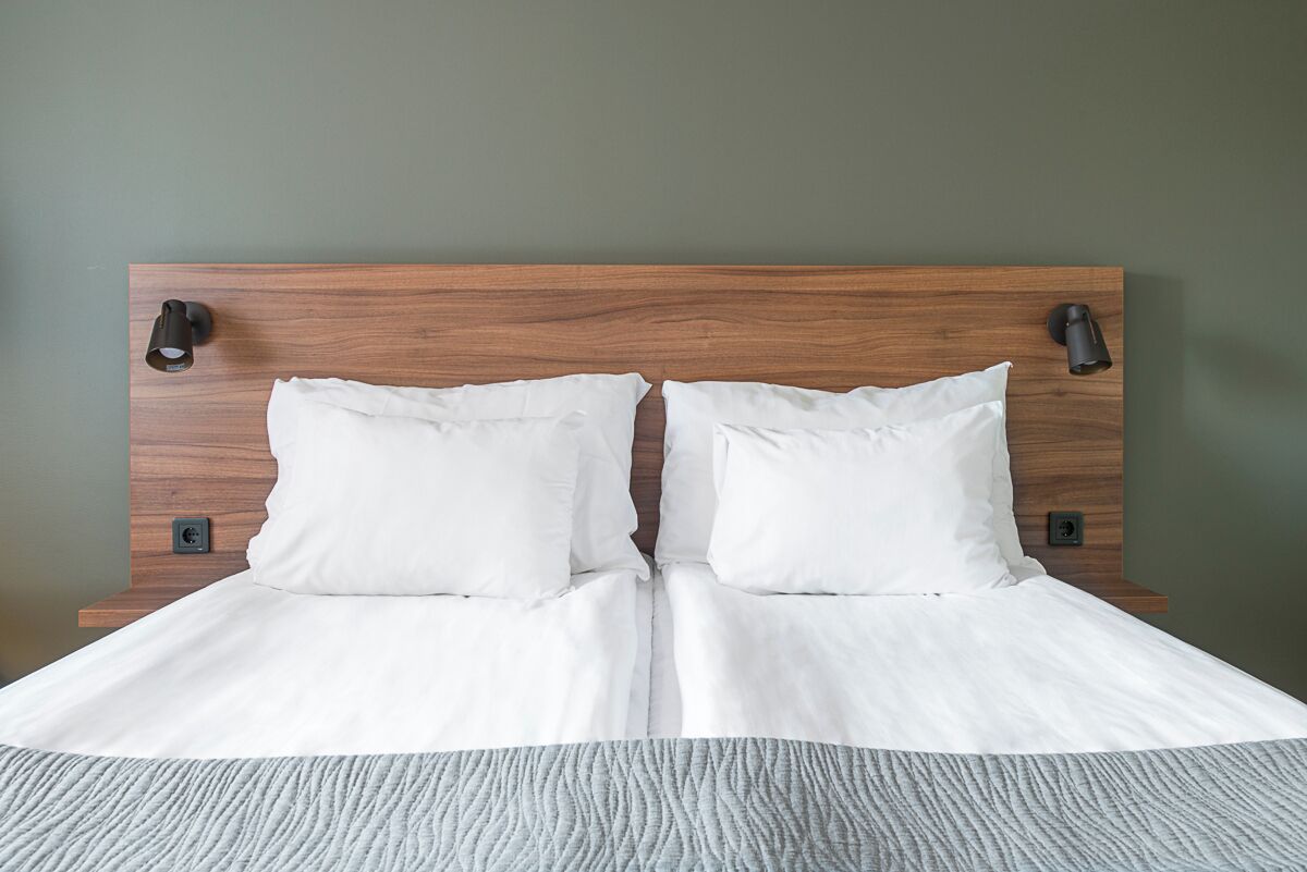 Een net opgemaakt bed met witte lakens en een grijs geweven sprei, geplaatst tegen een groene muur met een houten hoofdbord en twee wandlampen.