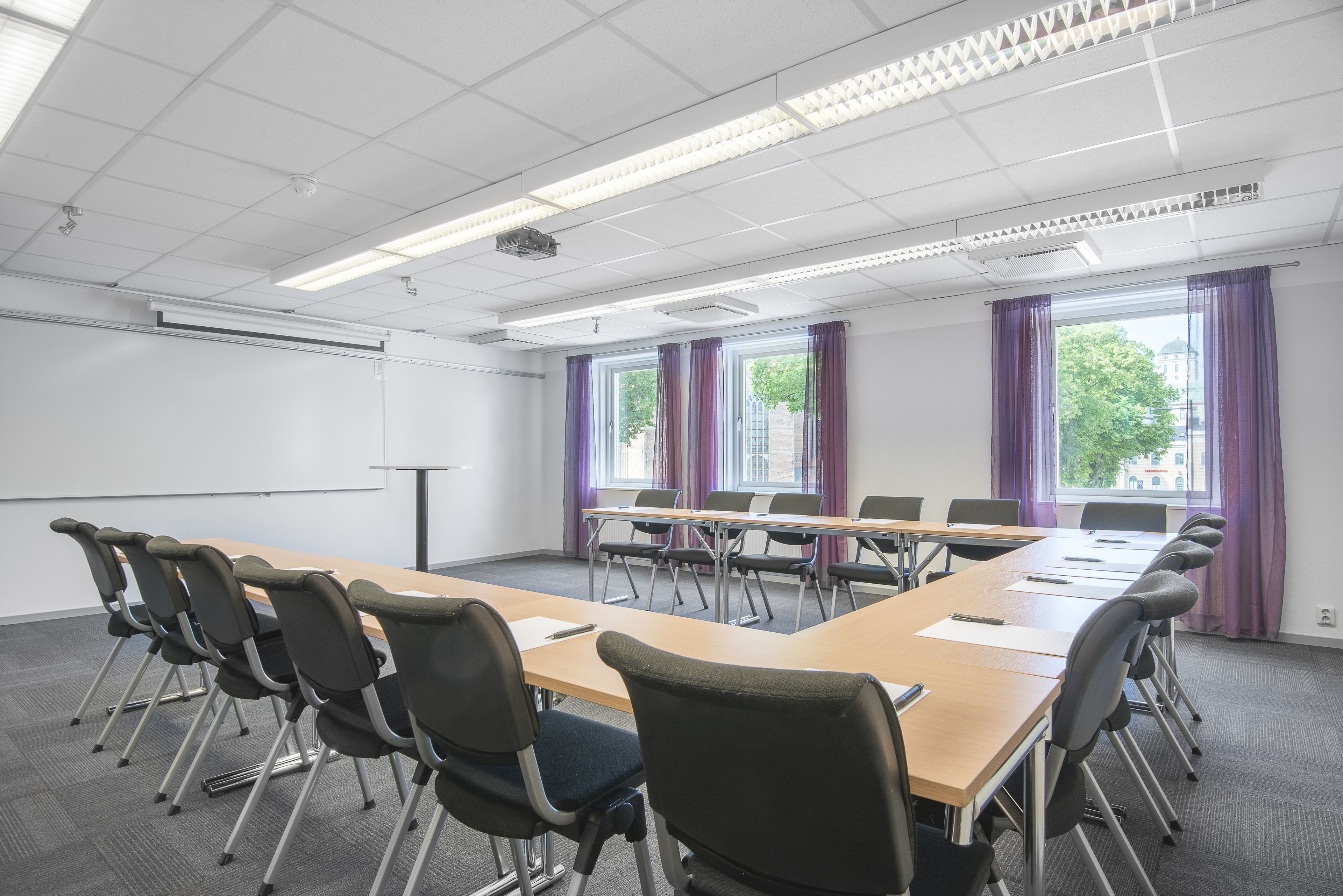 A conference room with a long table, chairs, a projector screen, and windows with curtains.