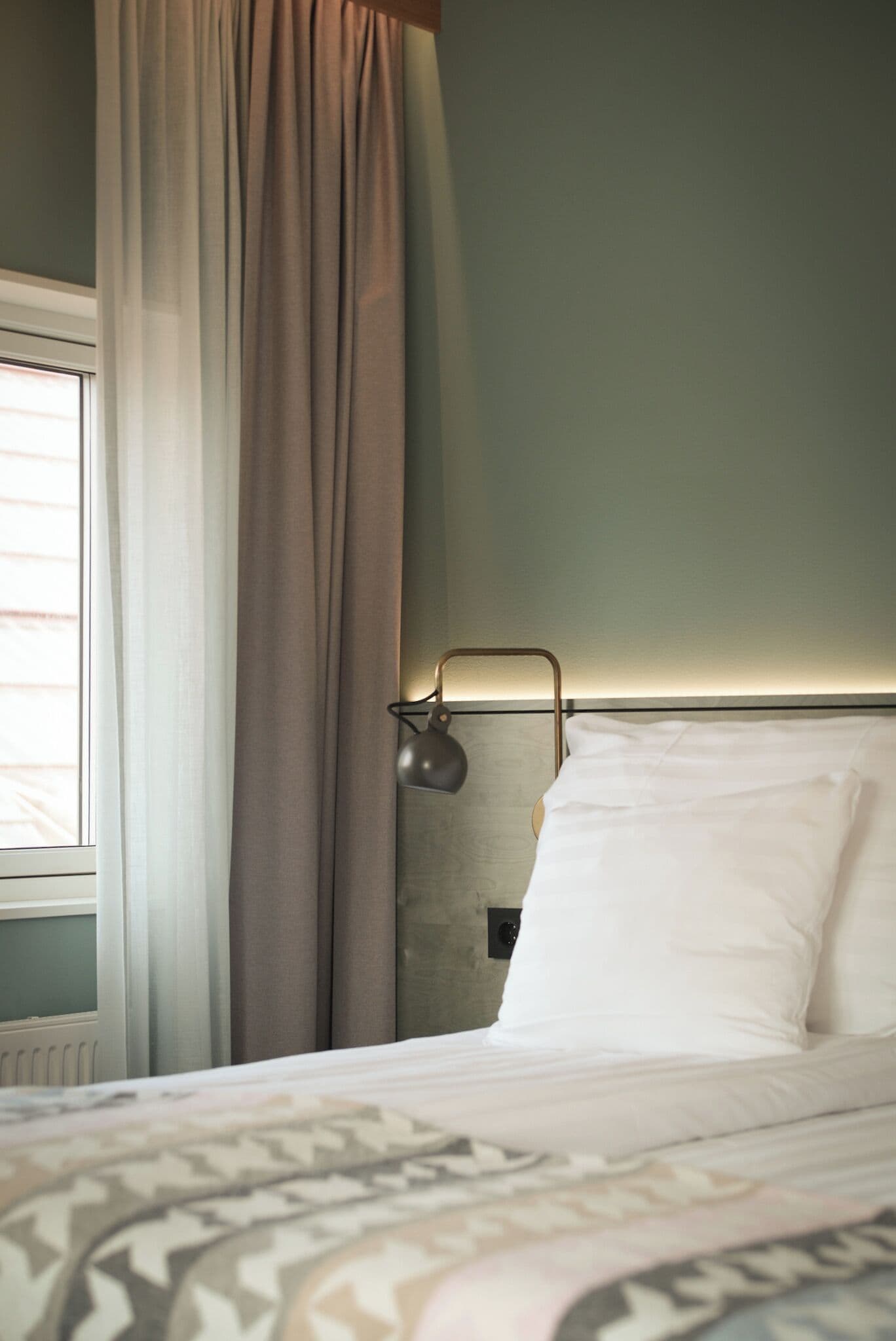 En pænt redt seng med hvide sengetæpper, en sengetavlelampe og et vindue med gardiner.