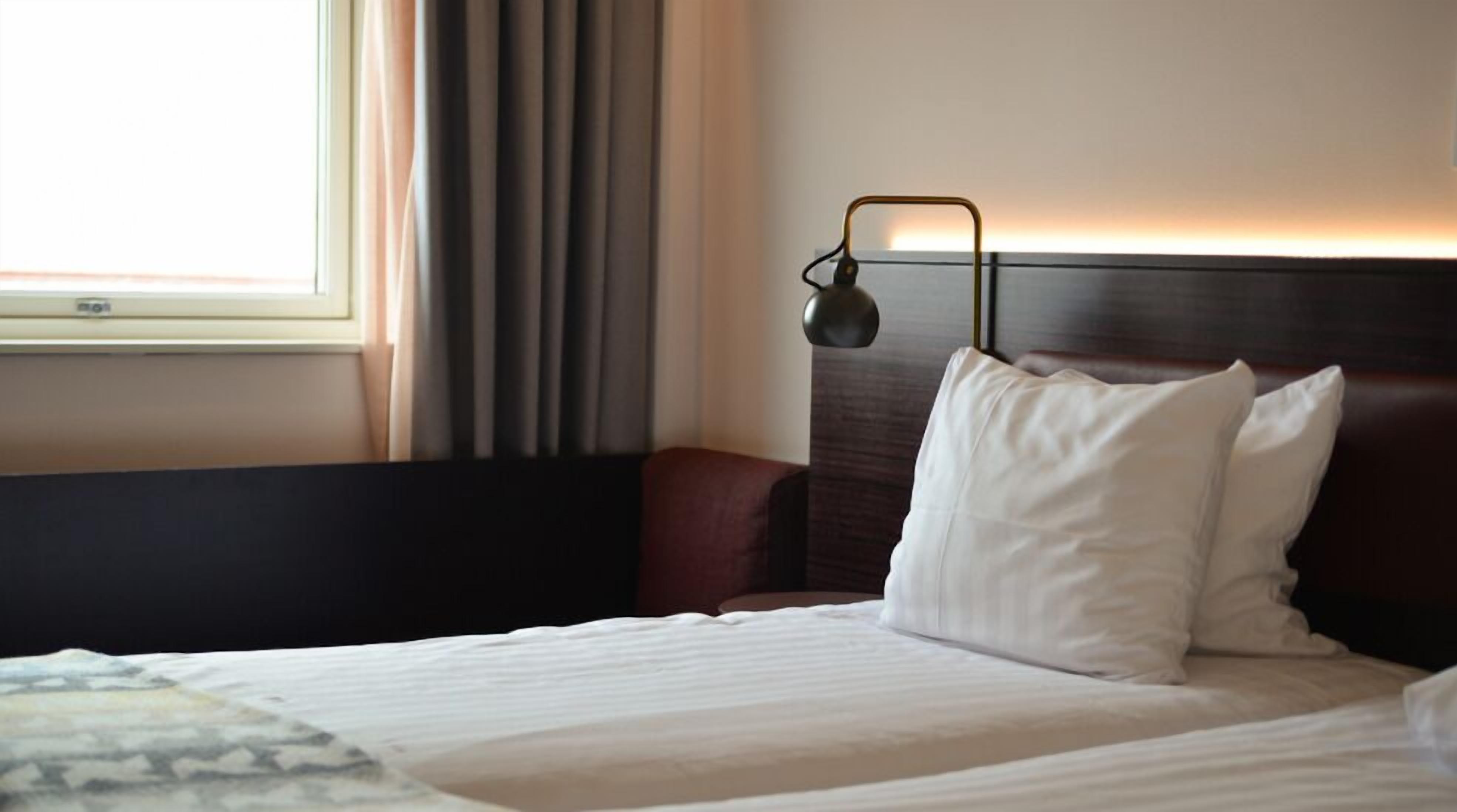 A hotel room with a bed, a nightstand, a lamp, and a window with curtains.