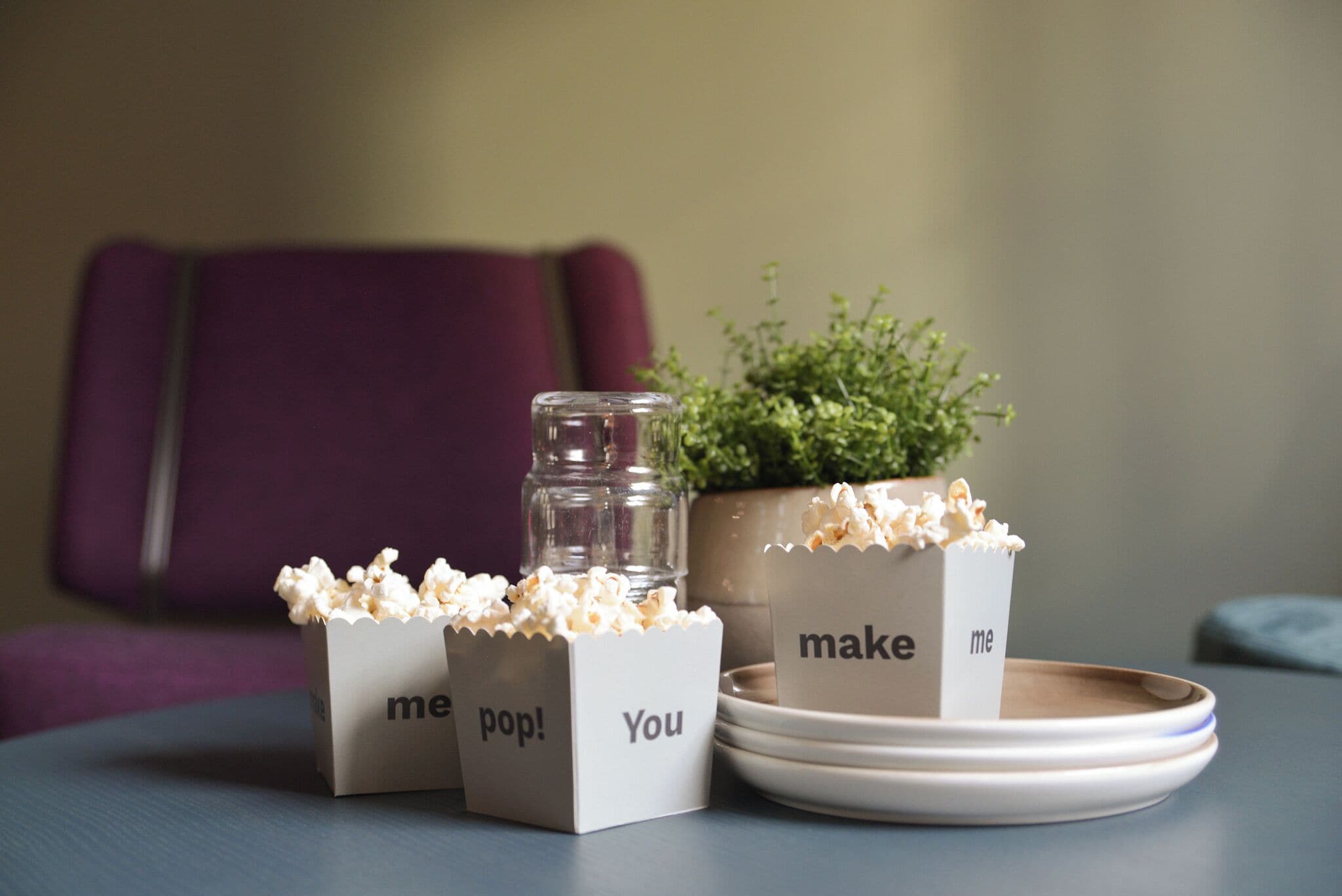 Tre popcorn beholdere med tekst, et glas og en potteplante på et bord.