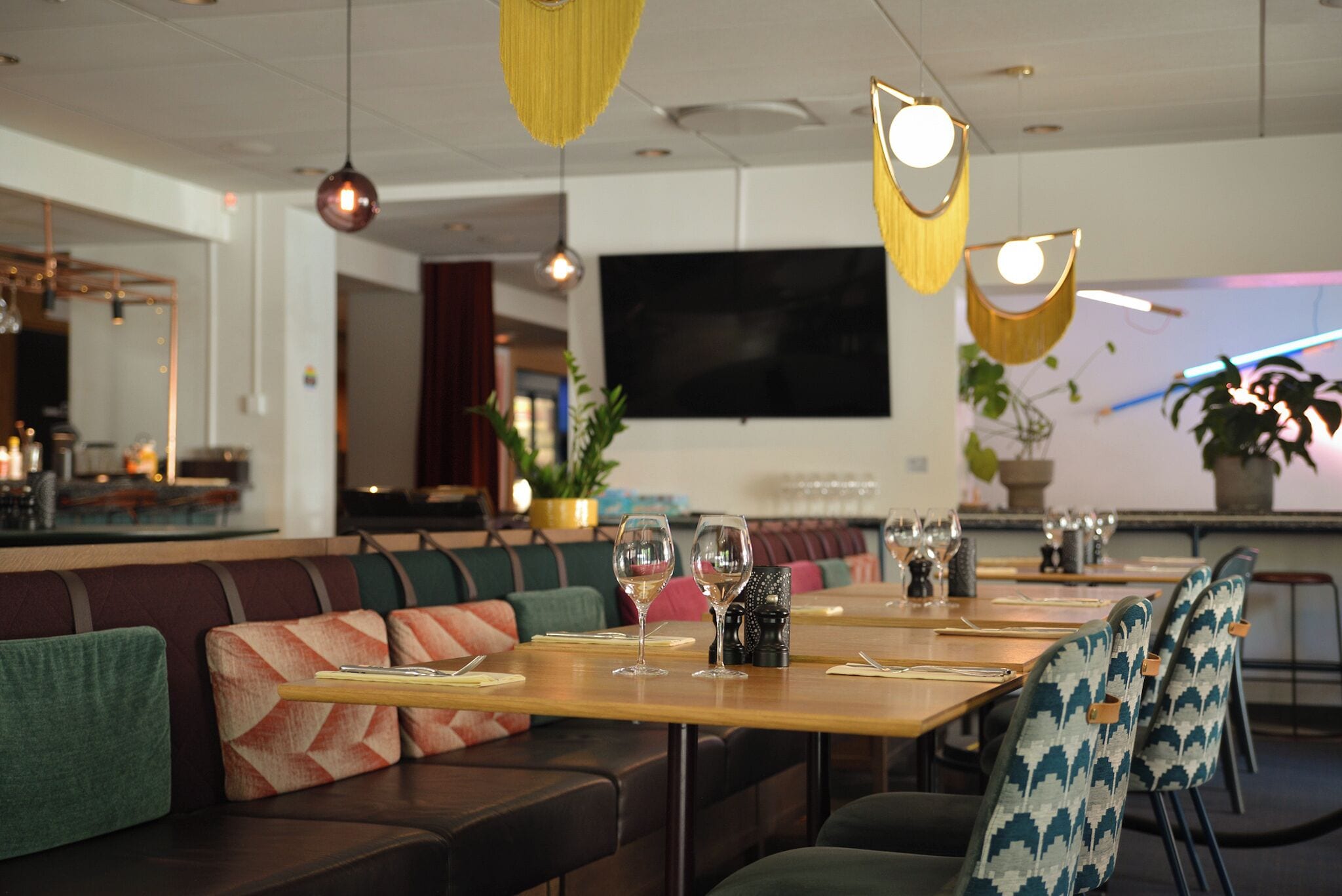A restaurant with a long wooden table set for dining, cushioned seating, hanging pendant lights, and a large flat-screen TV on the wall.