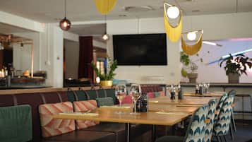 A restaurant with a long wooden table set for dining, cushioned seating, hanging pendant lights, and a large flat-screen TV on the wall.