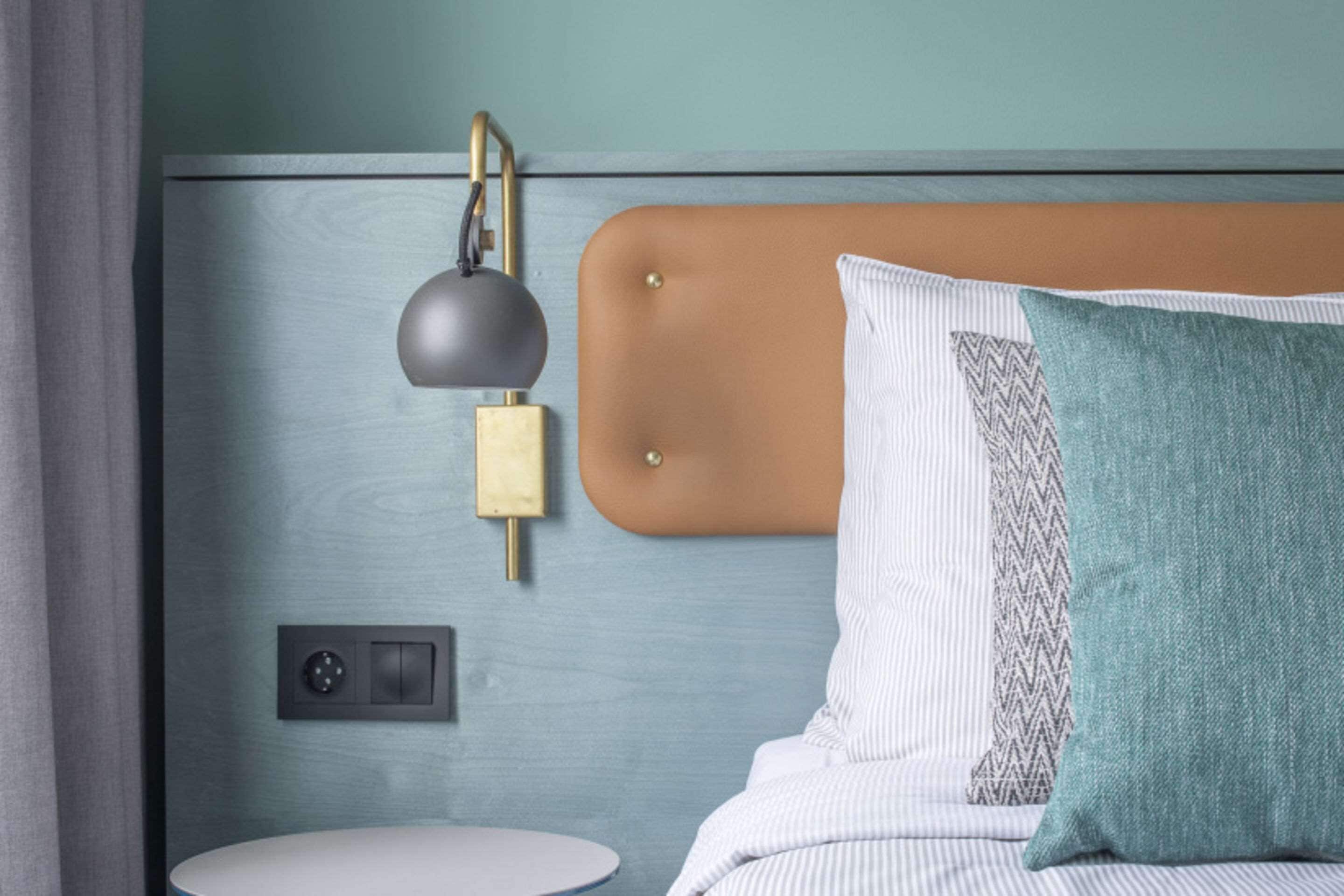 A bedroom with a bed, a bedside table, a wall-mounted lamp, and a control panel.
