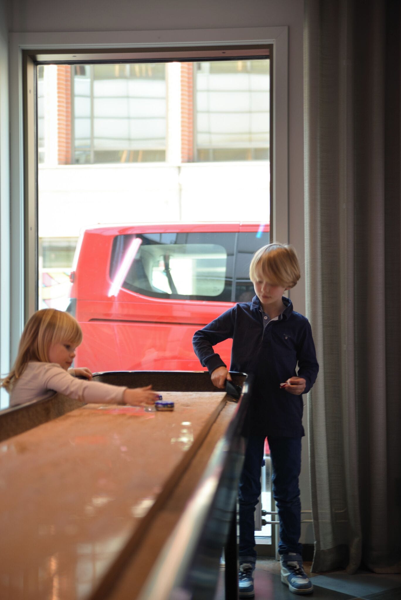 Dos niños en una mesa con vista a un vehículo rojo afuera.