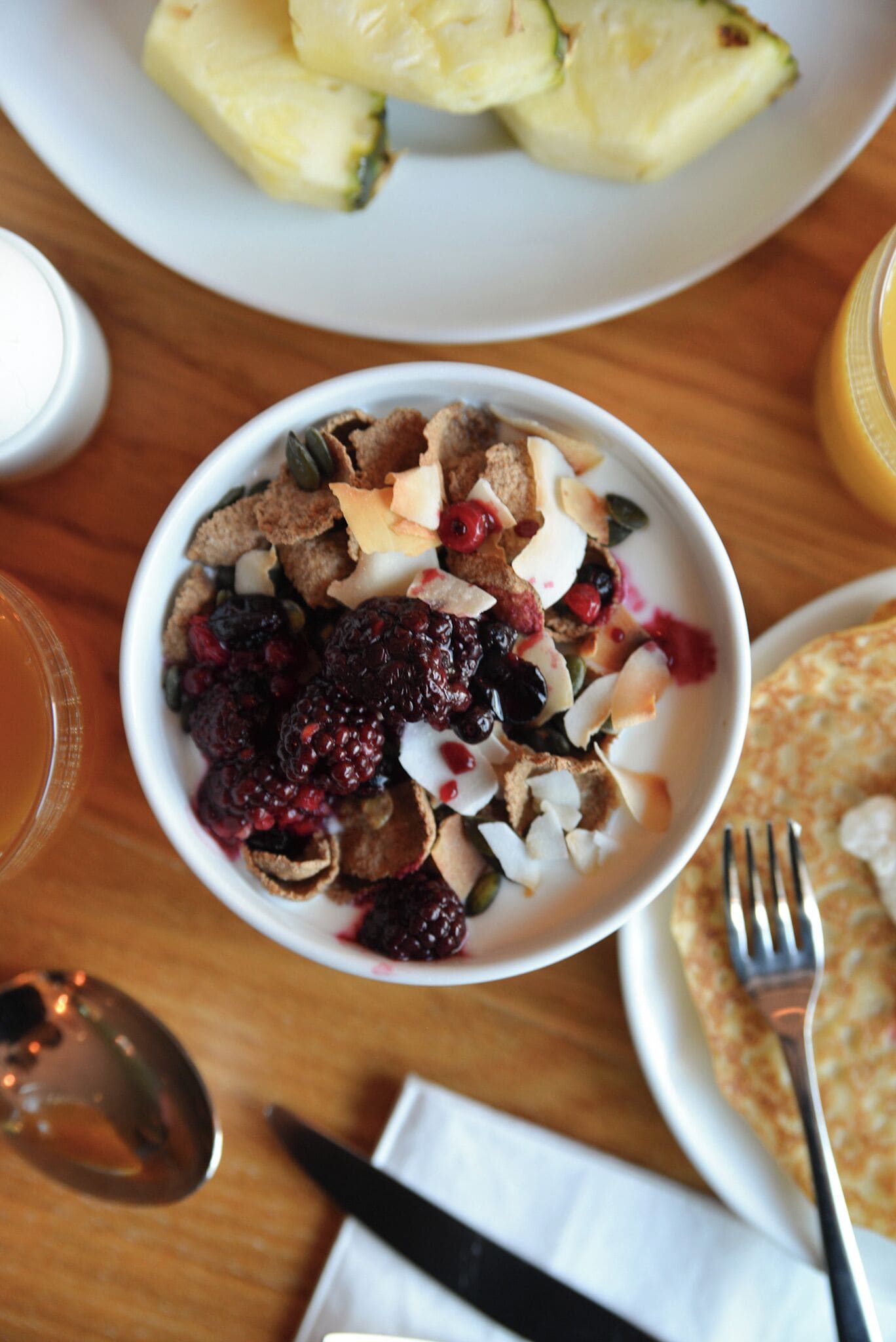 Un bol de céréales avec des fruits rouges et des noix, une assiette de crêpes et un verre de jus de fruits sur une table en bois.