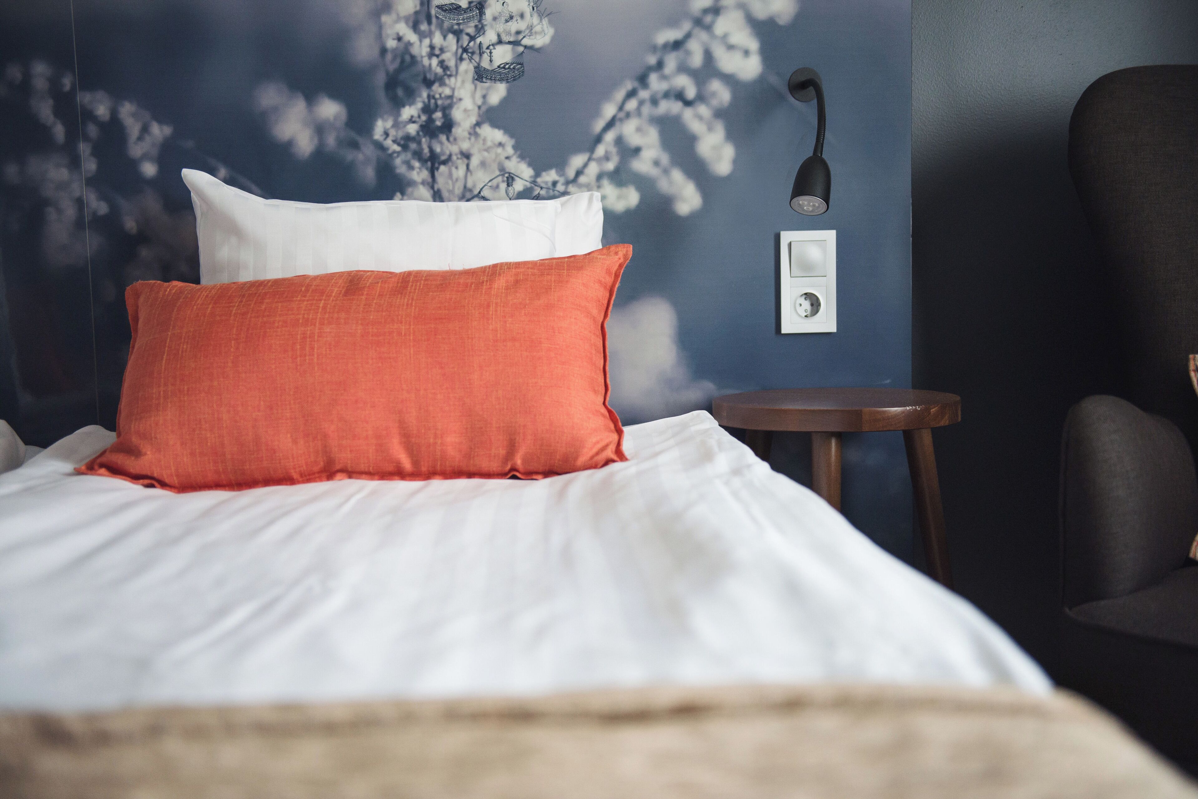 Een bed met witte en oranje kussens, een klein houten nachtkastje en een wandlamp.