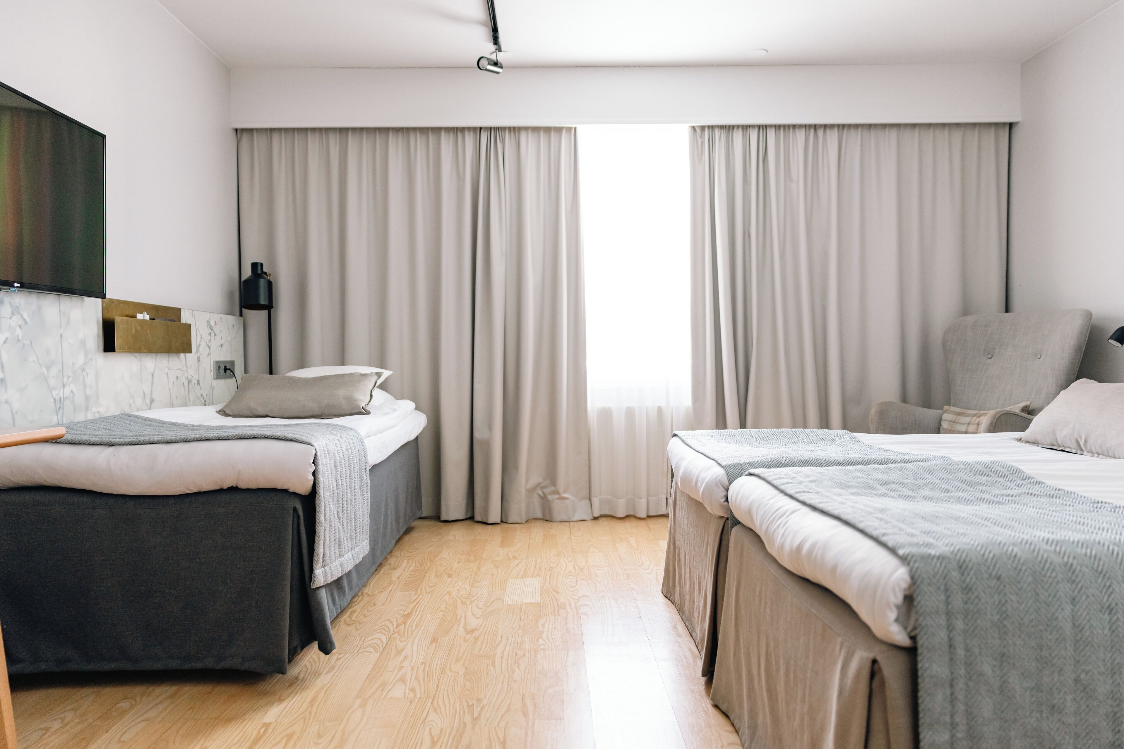 Ett hotellrum med två sängar, ett skrivbord, en stol, en TV och ett fönster med gardiner.