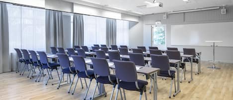Un salón de conferencias con hileras de sillas y mesas, ventanas amplias con cortinas, una pizarra y un proyector.