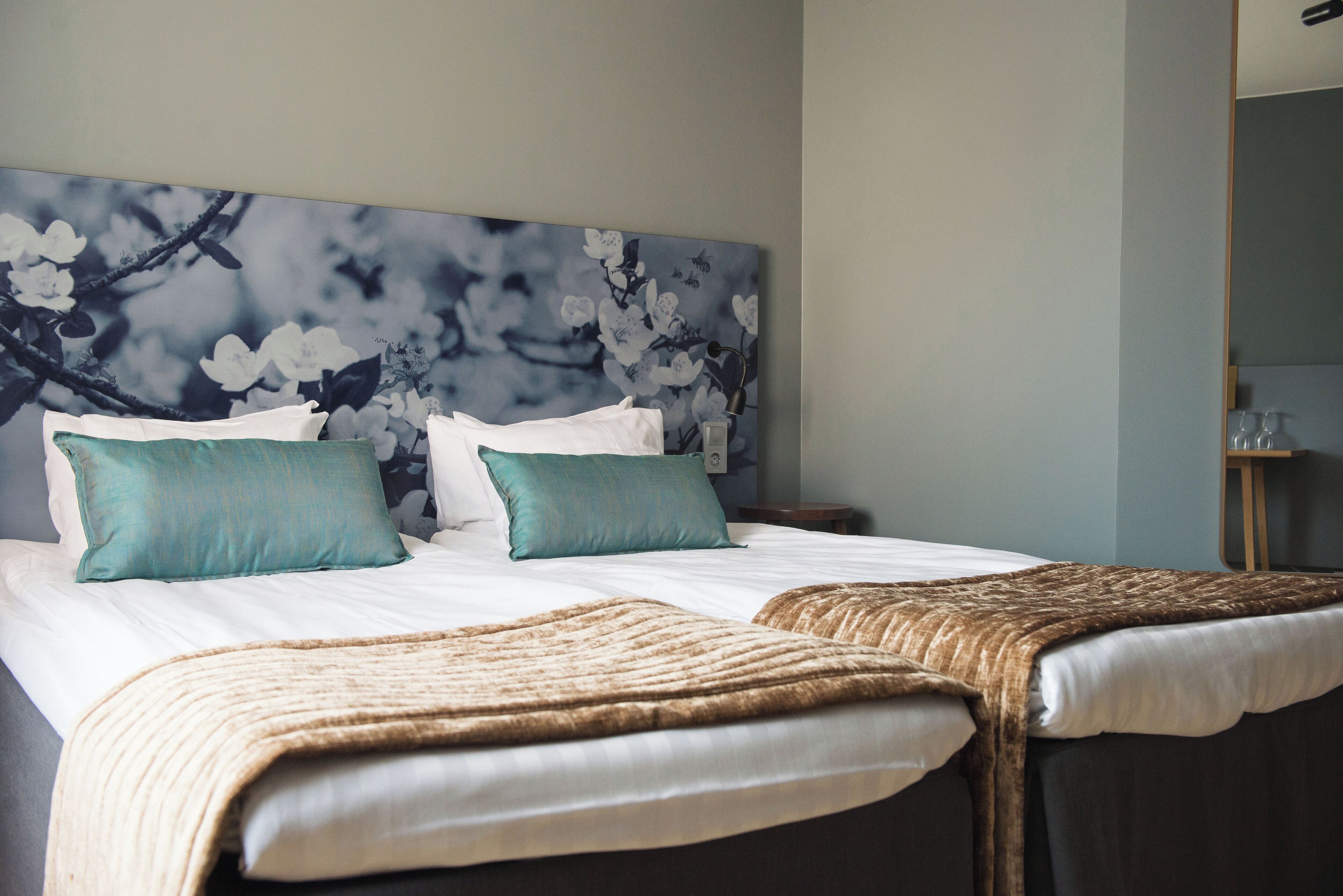 A hotel room with a large bed, two pillows, a headboard with a floral design, and a bedside table with a lamp.