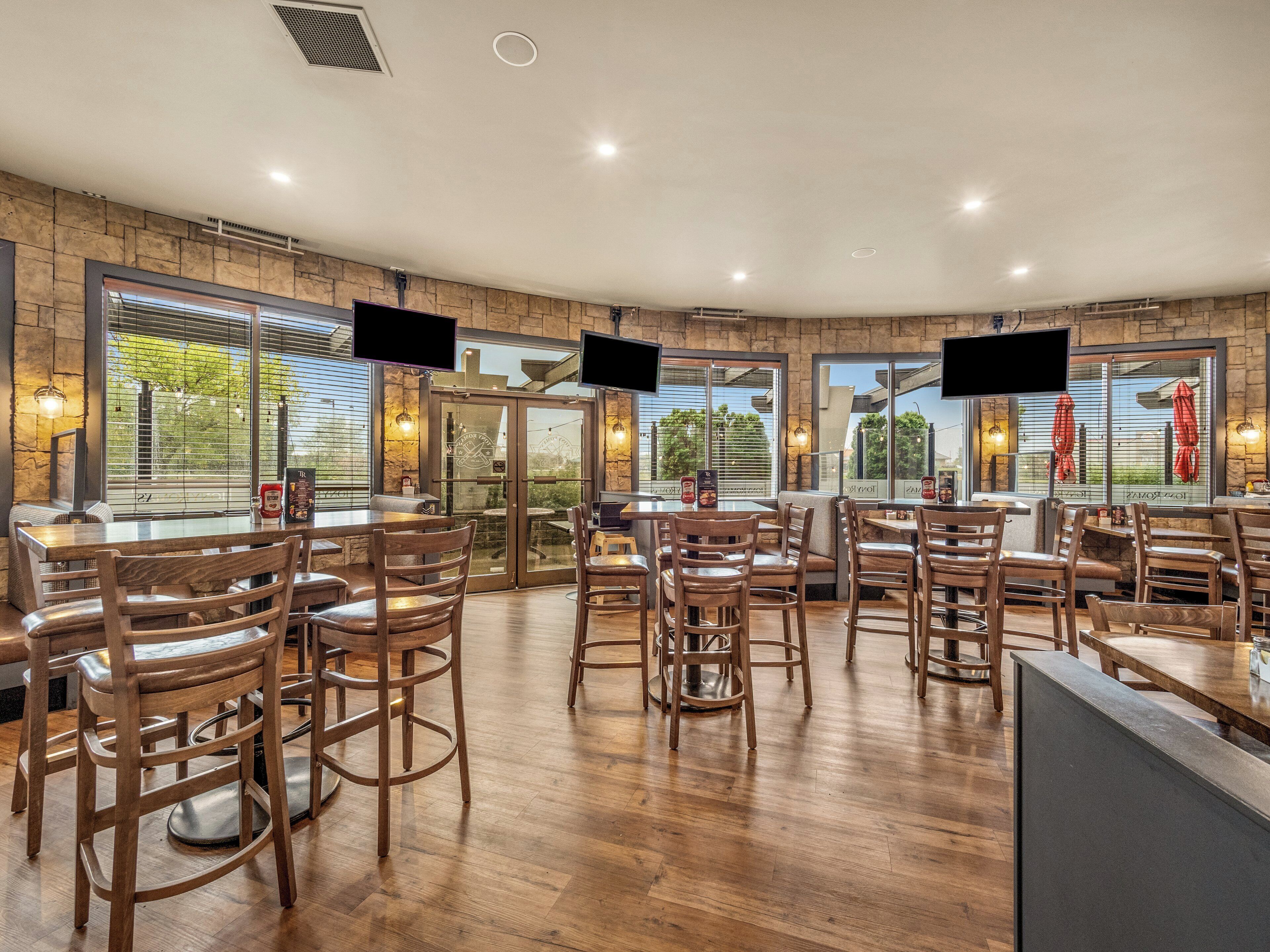 A restaurant with wooden flooring, multiple tables and chairs, large windows with blinds, and mounted TVs.