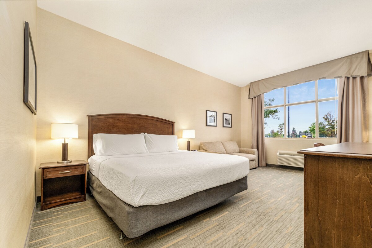 A hotel room with a large bed, a bedside table with a lamp, a sofa, a desk, and a window with curtains.
