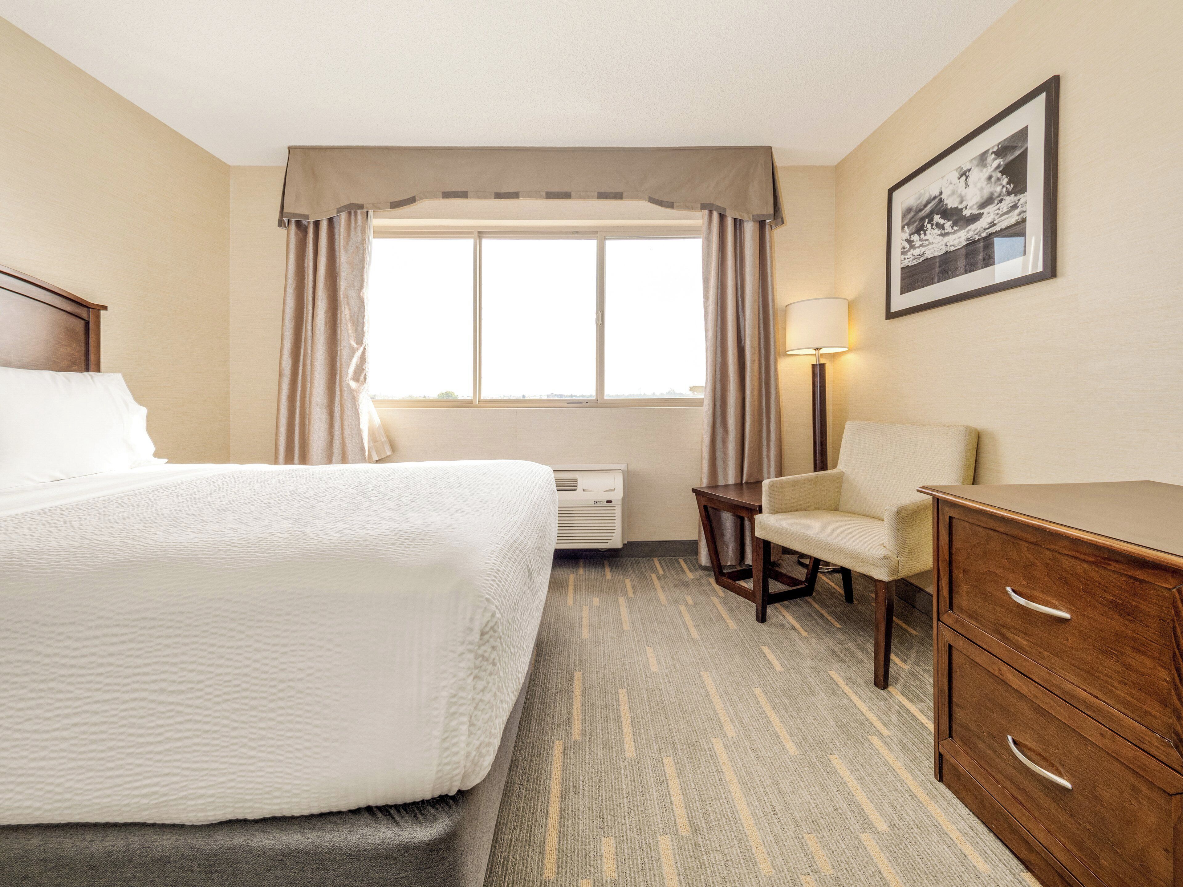A hotel room with a bed, a desk, a chair, a lamp, and a window with curtains.