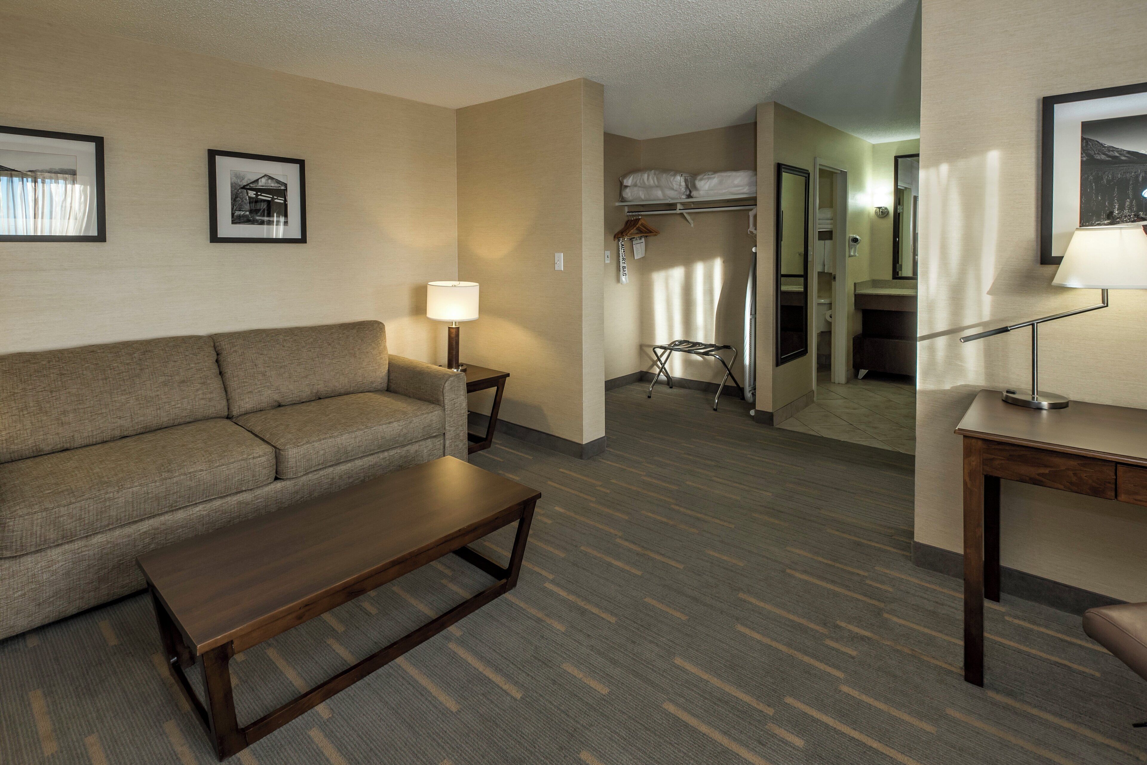 A hotel room with a sofa, a wooden coffee table, a lamp, a coat rack, and a closet with a bed.