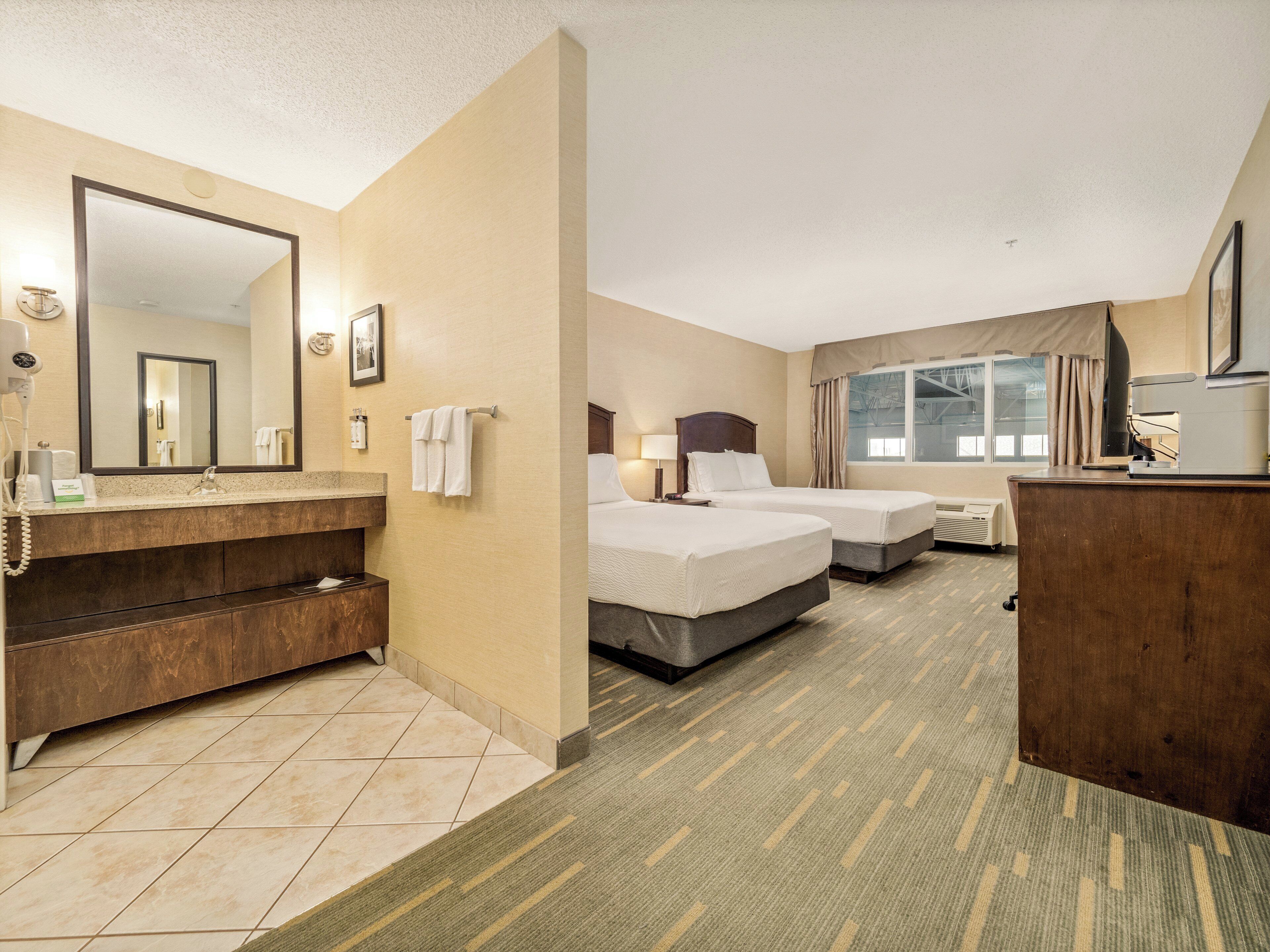 A hotel room with a bathroom, two beds, a desk, and a large mirror.