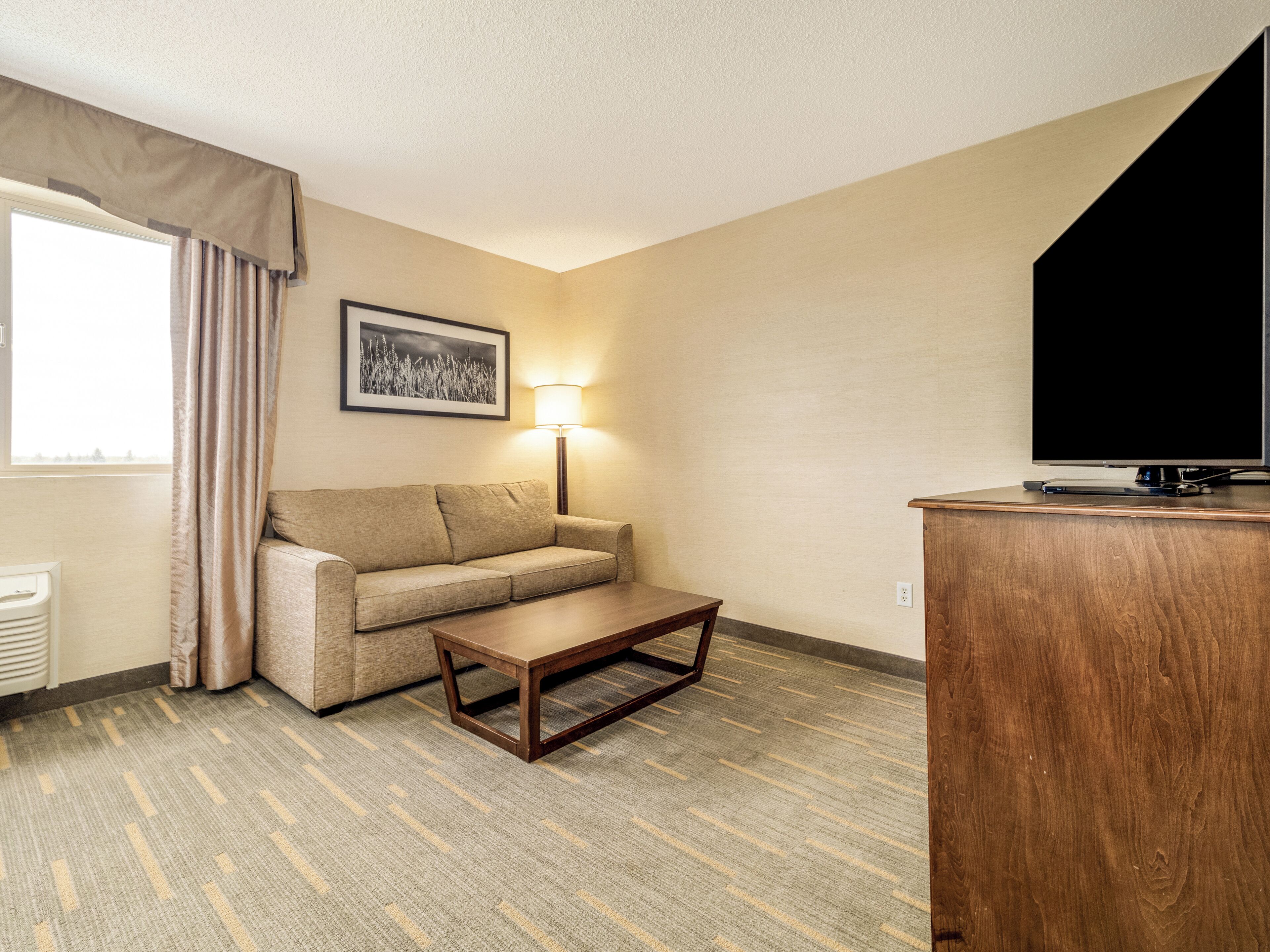 Une chambre d’hôtel avec un canapé, une table basse, une lampe, un téléviseur et une fenêtre avec des rideaux.