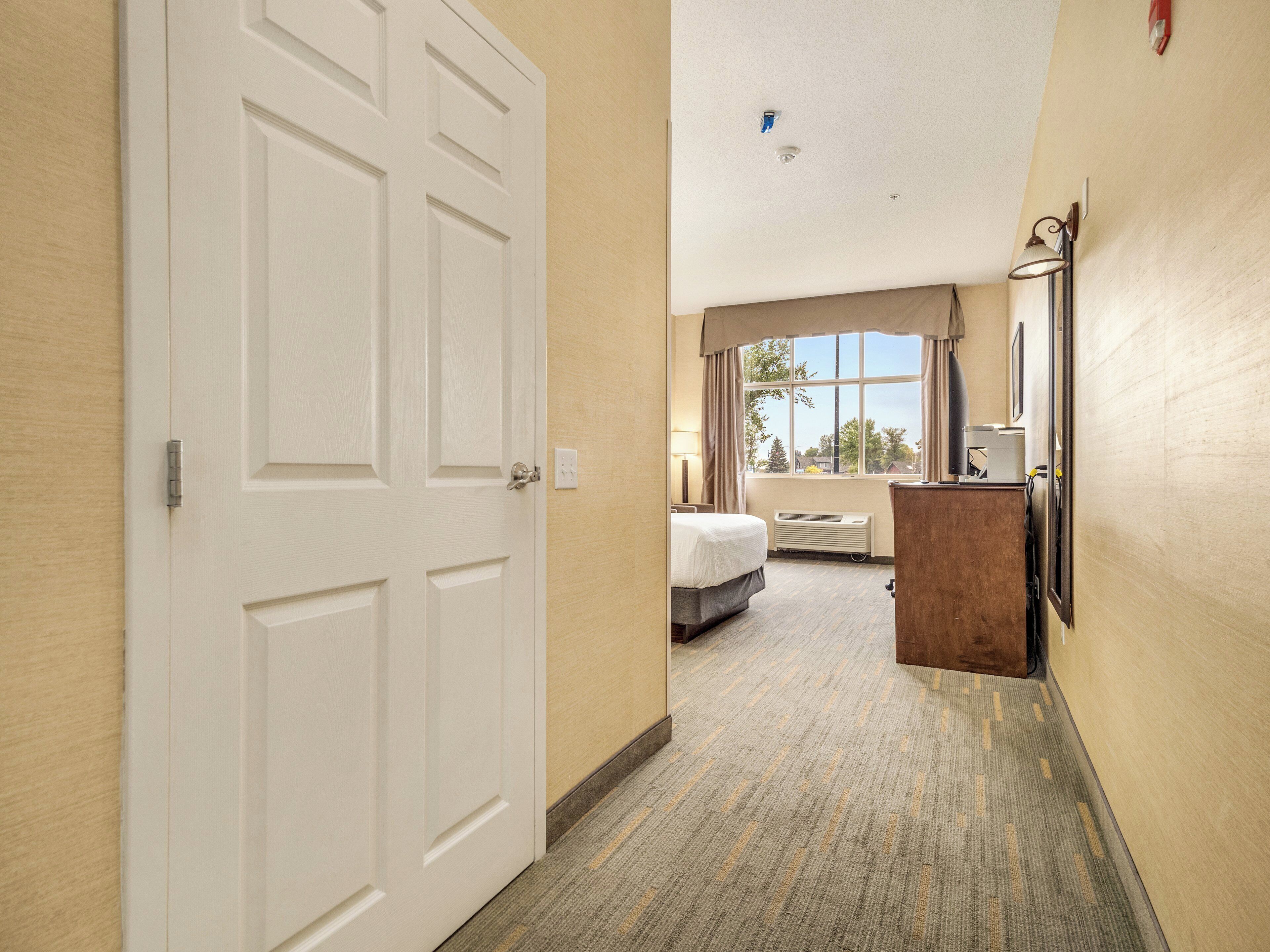 A hotel hallway leading to a bedroom with a bed, a dresser, and a window with a view.
