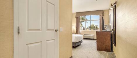 A hotel hallway leading to a bedroom with a bed, a dresser, and a window with a view.