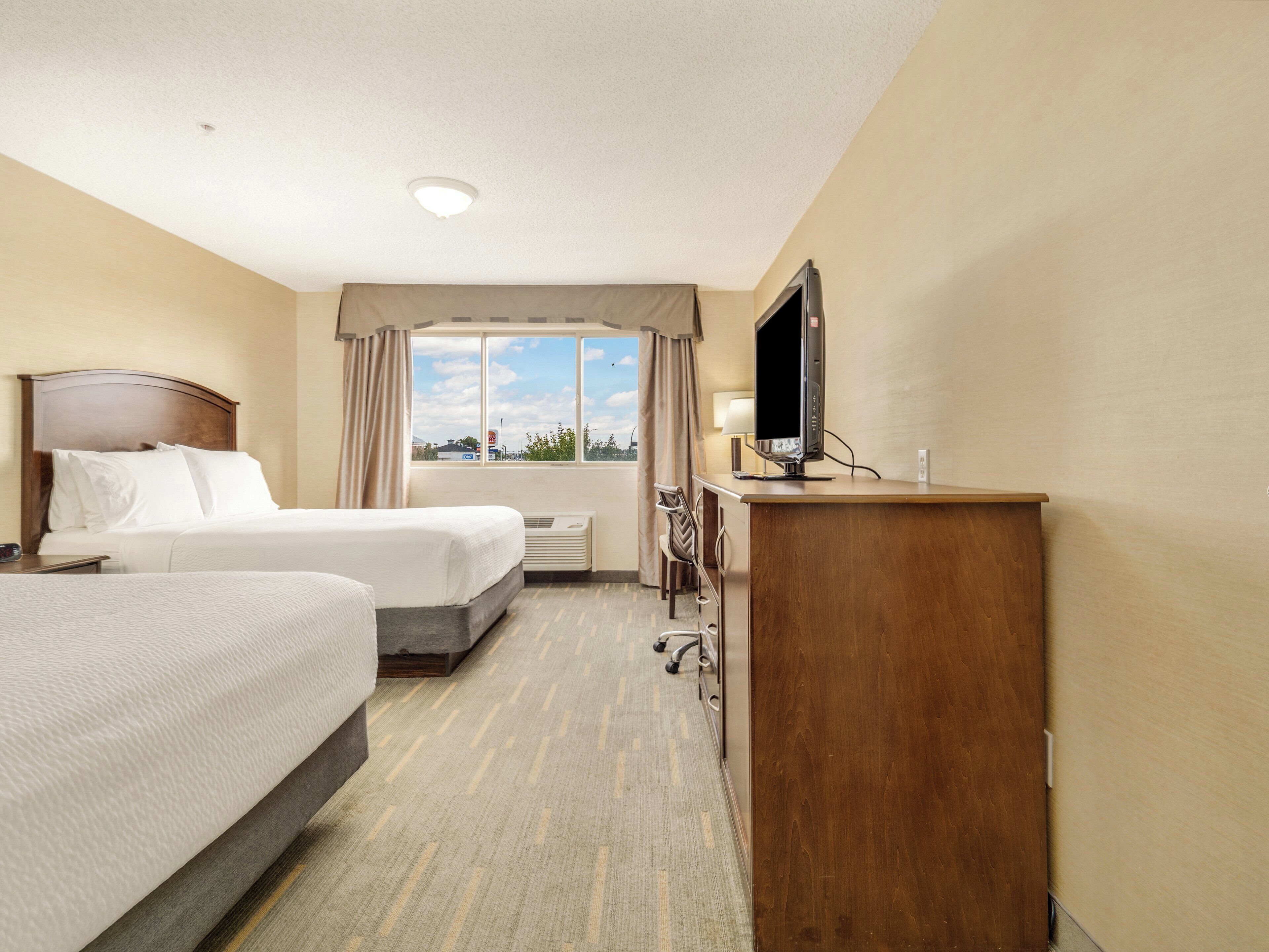 A hotel room with two beds, a desk with a TV, a chair, and a window with a view of buildings.