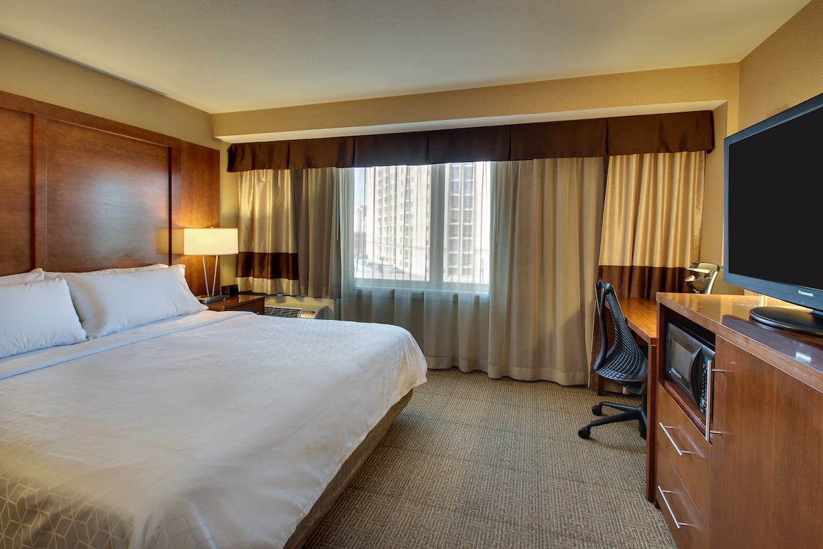 A hotel room with a large bed, a desk with a television, a chair, and a window with curtains.