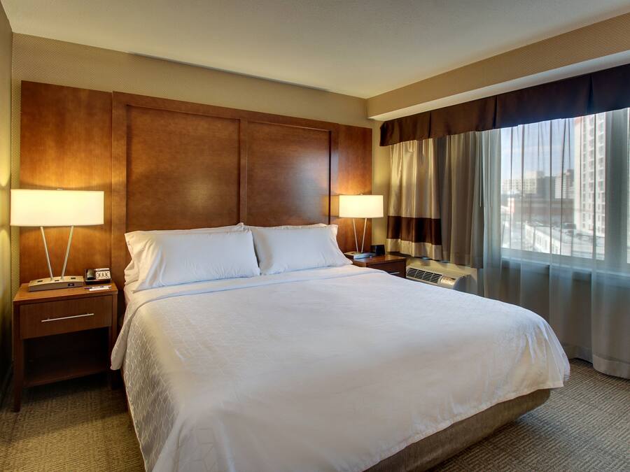 Ein Hotelzimmer mit einem großen Bett, zwei Nachttischen, einer Lampe und Blick auf die Stadt durch das Fenster.