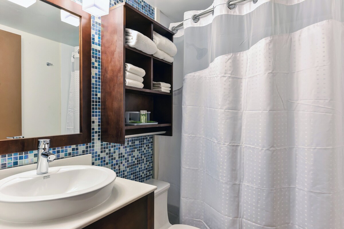 A bathroom with a white sink, a mirror, a shower curtain, and a shelf with folded towels.